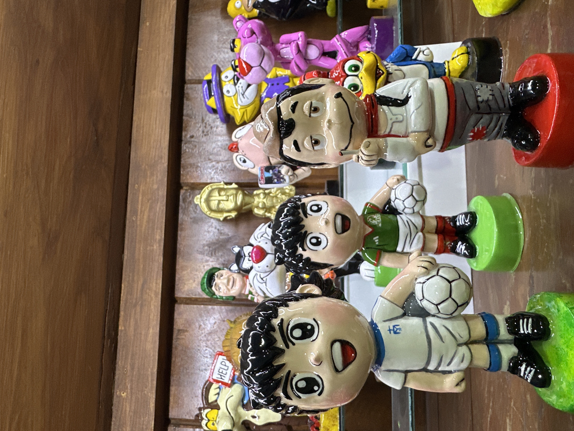 The image shows several painted ceramic figurines, likely in a retail setting. The figurines depict cartoon characters, including soccer players, and other figures. The soccer players wear different colored jerseys, one with a white jersey and blue trim, and another with a green jersey. The figurines are displayed on a shelf with a wooden background. Based on the style of the figurines and their presence in the city of Mexico City, Mexico, it is possible they are souvenirs or collectibles popular in the region. The specific location cannot be determined solely from the image.
