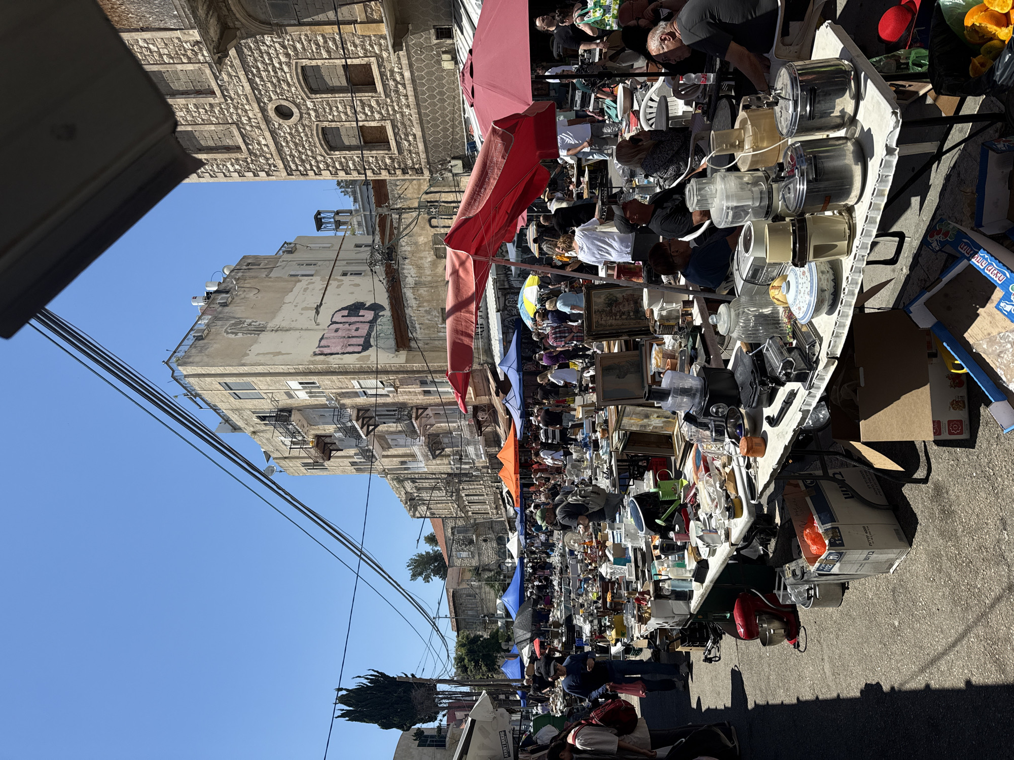 This is an outdoor scene of a crowded flea market set up on a street in Kiryat Ata, Israel. The market stalls are lined with a variety of second-hand items, including kitchen appliances, household goods, and other miscellaneous objects. People are browsing the stalls, examining items for sale. Red, blue, and orange umbrellas provide shade over the market. Buildings line the street on either side of the market. Above the buildings, a clear blue sky is visible.