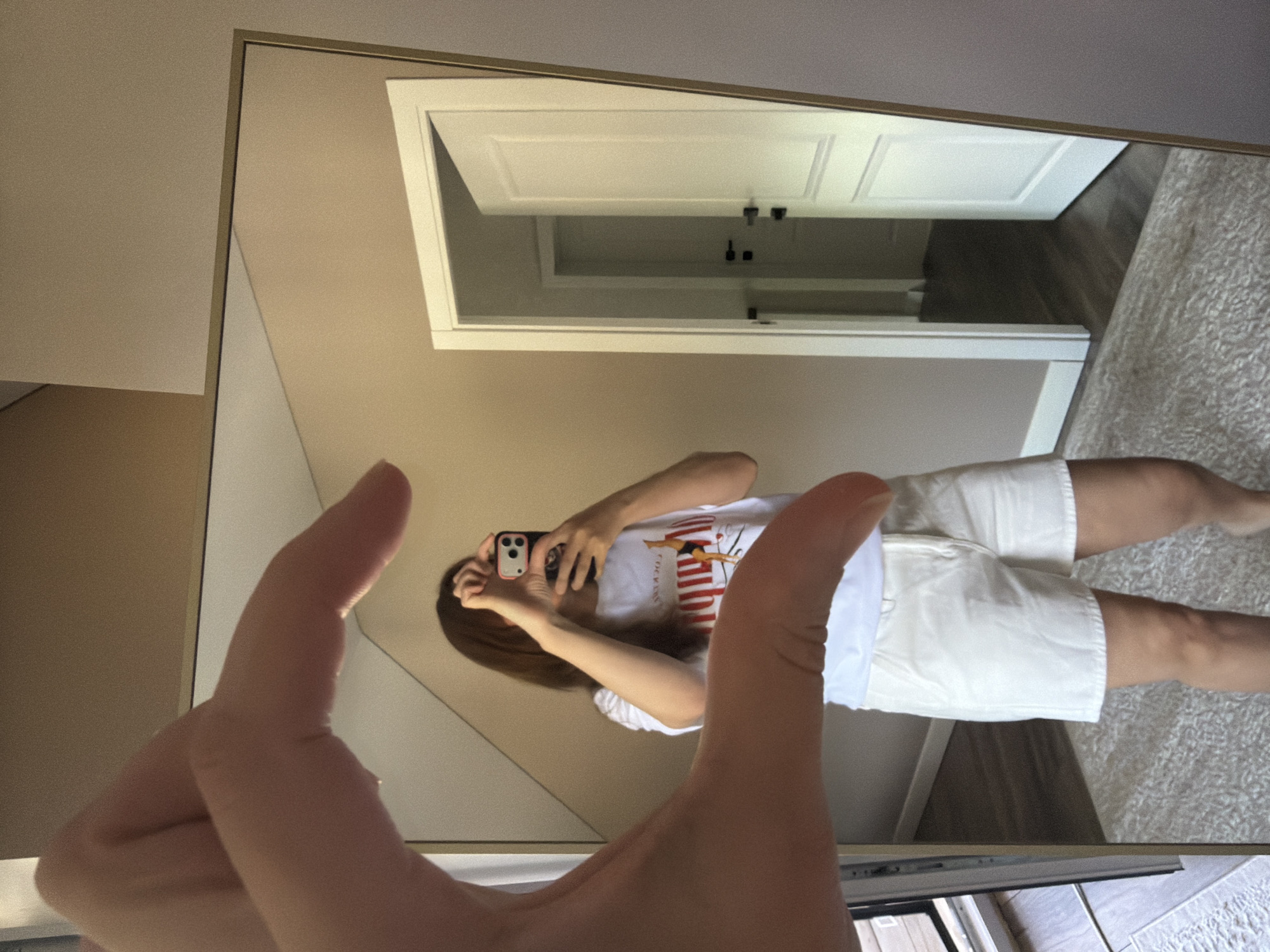 A person, reflected in a full-length mirror, is holding a smartphone and taking a selfie. They are wearing a white t-shirt with a graphic print and white shorts. The background shows a doorway with a white door ajar, leading to another doorway in the distance. The room has light-colored walls and a textured rug on the floor. A large, golden-framed mirror dominates the foreground, with the photographer's hand creating a heart shape in the foreground, obscuring part of the reflection. The scene is indoors, in what appears to be a residential setting.