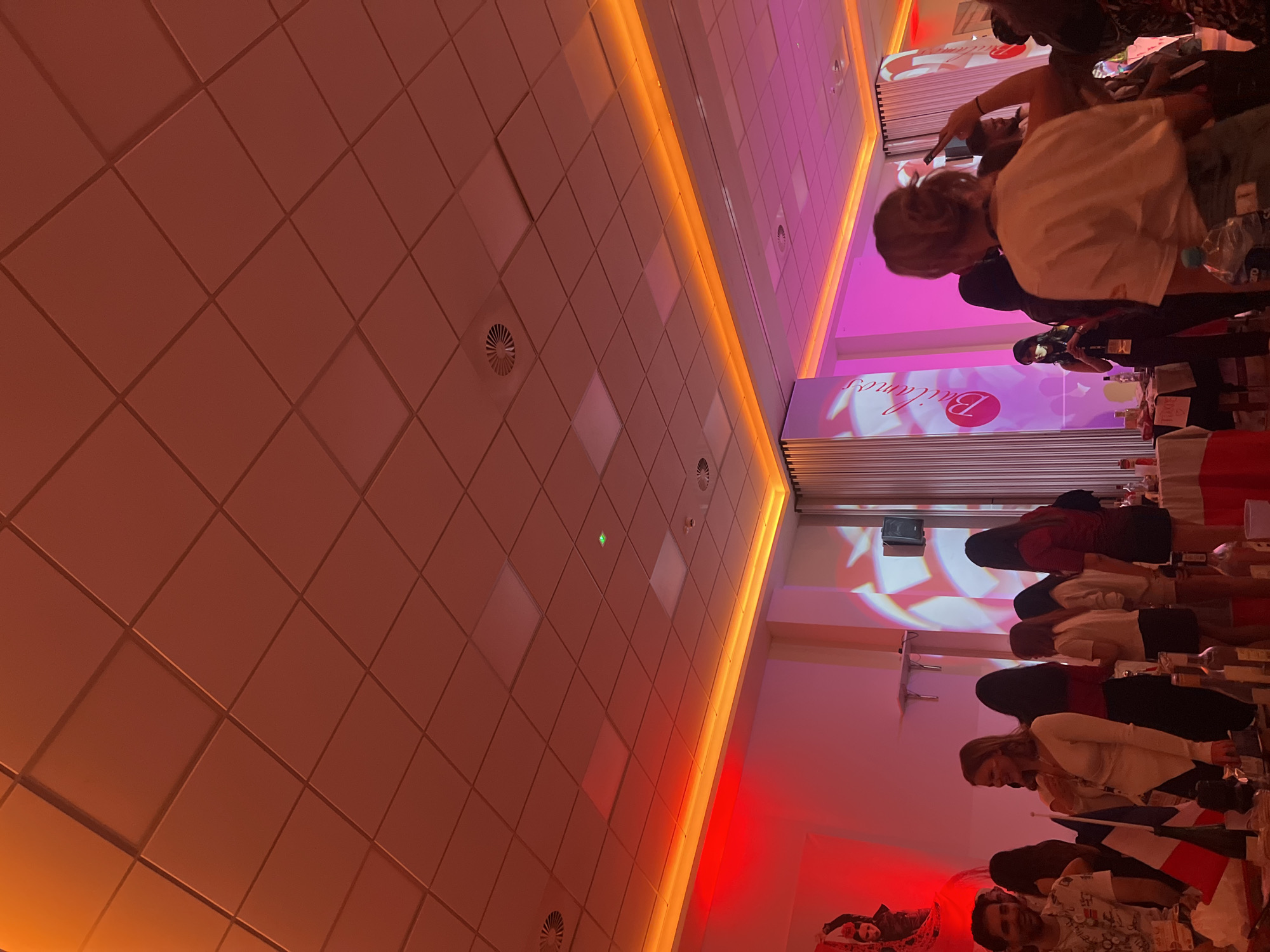 The image depicts an indoor event or gathering in what appears to be a modern hall, viewed from a slightly elevated and rotated perspective. The ceiling, dominating the left side of the frame, is covered with light-colored, square grid tiles, some with subtle texture or embossing, and includes several circular air vents. Strips of warm orange LED lighting illuminate the ceiling edges, creating a dynamic glow.
On the right side of the image, numerous individuals, primarily women, are gathered, standing in rows. They appear to be attentive, possibly watching a presentation or interacting. Their attire is varied, ranging from casual to semi-formal. In the foreground, small flags, including a clearly visible French flag, are placed on tables covered with red and white cloths. Bottles, possibly of water or other beverages, are also visible on these tables. A man with a beard is noticeable in the lower right corner, looking towards the camera.
A large banner or screen in the background is brightly lit with pink and magenta uplighting, displaying the word "Brilliance" (or "Brillance") in a elegant, cursive font, along with a stylized 'B' logo. Additional red spotlights cast a vibrant glow on the lower walls and floor areas. The combination of varied artificial lighting (orange, pink, magenta, red) creates a lively and energetic atmosphere, suggesting an evening event or a venue designed for presentations and social interaction in Bydgoszcz, Poland.