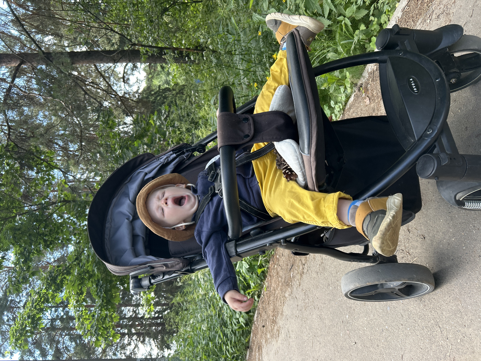 A young child in a stroller is yawning. The child is wearing a tan bucket hat, a navy blue long-sleeved shirt, and yellow pants. The stroller is black with a brown handle. The child is sitting in the stroller in a park with trees and green foliage in the background. The ground is a paved path. This appears to be an outdoor scene in the city of Chaikovsky, Russia.