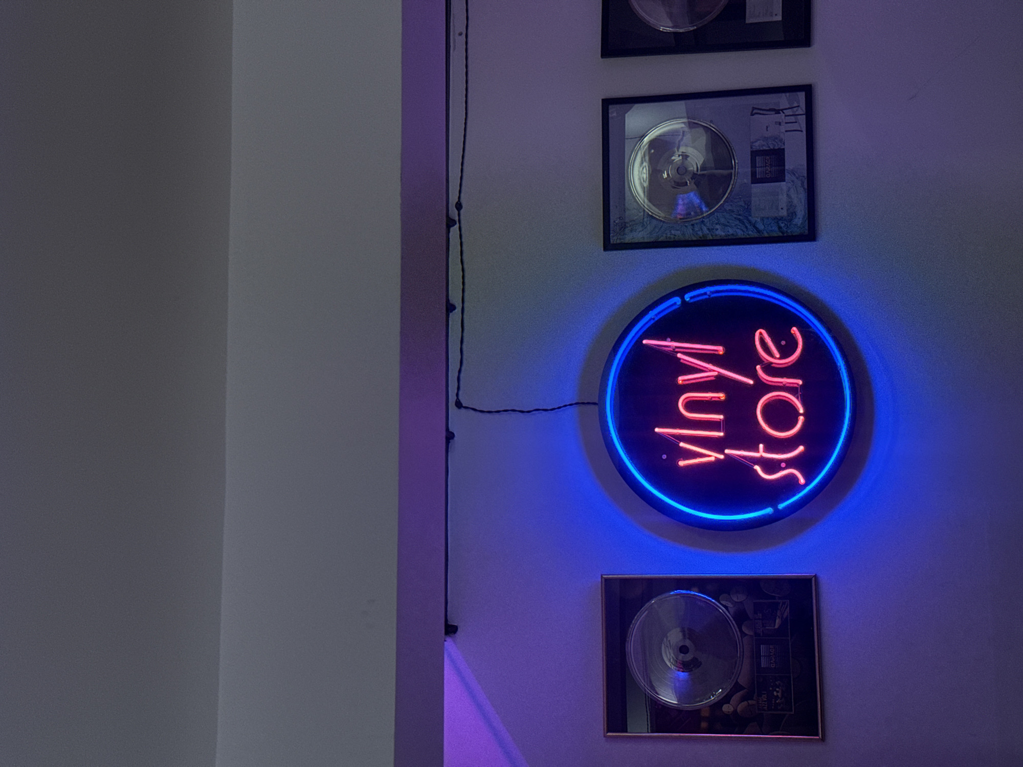 The image shows a wall with decorations in an interior setting. A neon sign that reads "vinyl store" is prominently displayed, framed by a blue neon circle. Above and below the sign are framed objects, possibly vinyl records or other memorabilia. A vertical cable runs alongside a wall edge, from the top to the bottom of the image. The lighting is dim, creating a blue and purple hue. There is no information to place this image within Yerevan, Armenia.