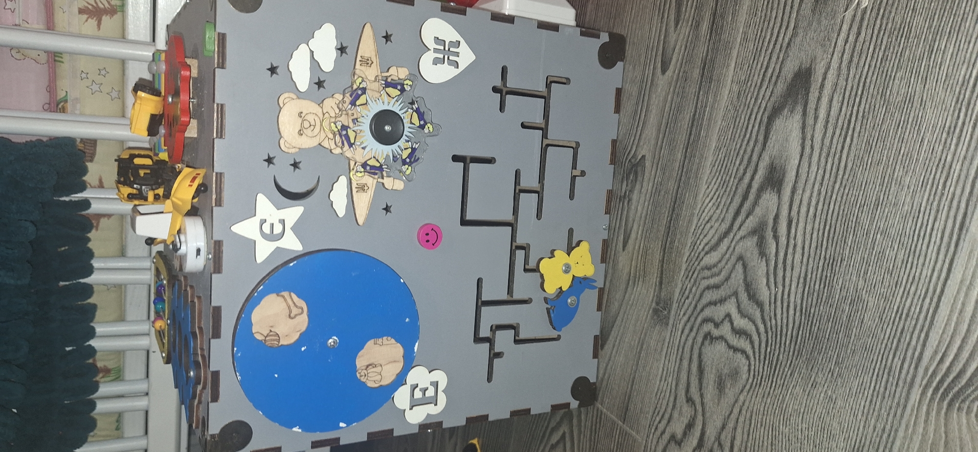The image shows a grey wooden sensory cube toy placed on a grey wooden floor. The toy has various colorful elements on its sides, including a blue disc with images of planets, a white star with the letter "E" inside, a cartoonish teddy bear riding a plane, a pink smiley face, and a maze pattern with two yellow and blue animal figures. The background appears to be a crib with green and white elements. The toy is positioned indoors. There is no indication of the city of Bila Tserkva, Ukraine, in the image.