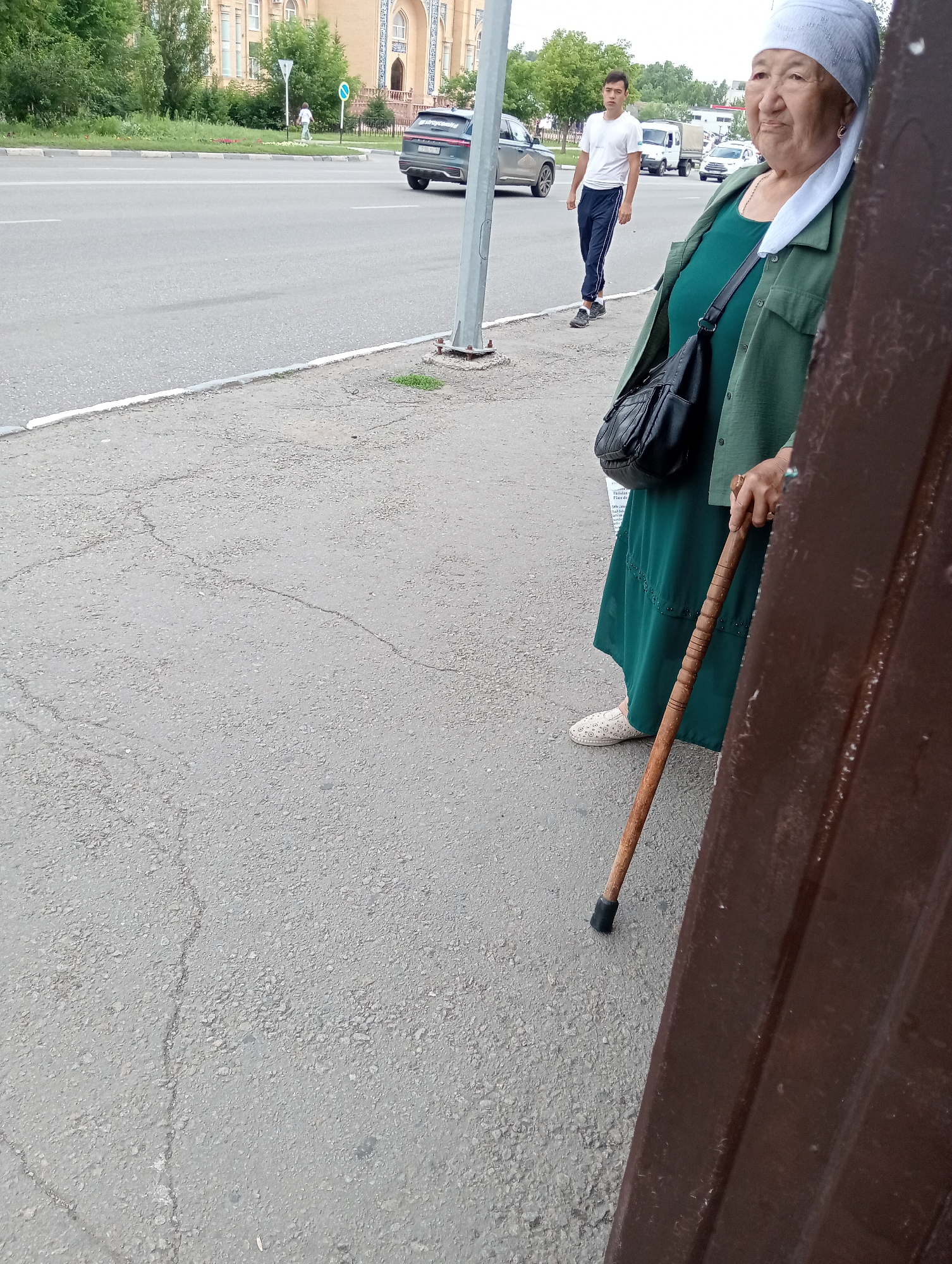 An older woman wearing a white headscarf and a green outfit stands on a cracked asphalt sidewalk, leaning on a wooden cane. She has a black purse slung over her shoulder. In the background, a young man in a white t-shirt and dark pants walks along the sidewalk. Several cars are visible on the road, and a light-colored building with arched windows and decorative details stands behind a grassy area.