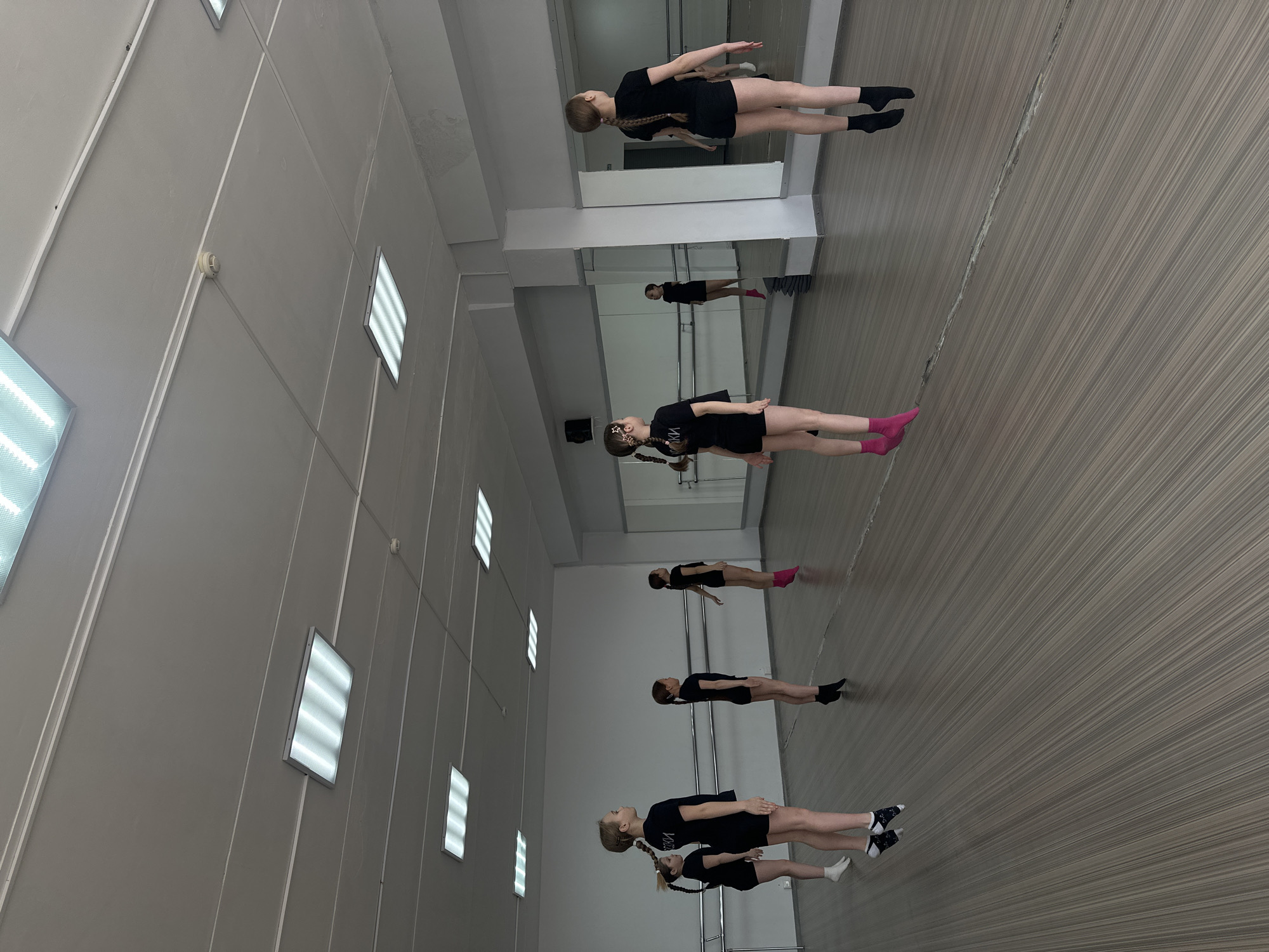 The image shows a dance studio. Six girls in black shirts and shorts are lined up in a row, practicing poses. The studio has a wall of mirrors, and the floor has a striped wood pattern. The room is lit by fluorescent lights on the ceiling. The location is likely Votkinsk, Russia, given the prompt's request.