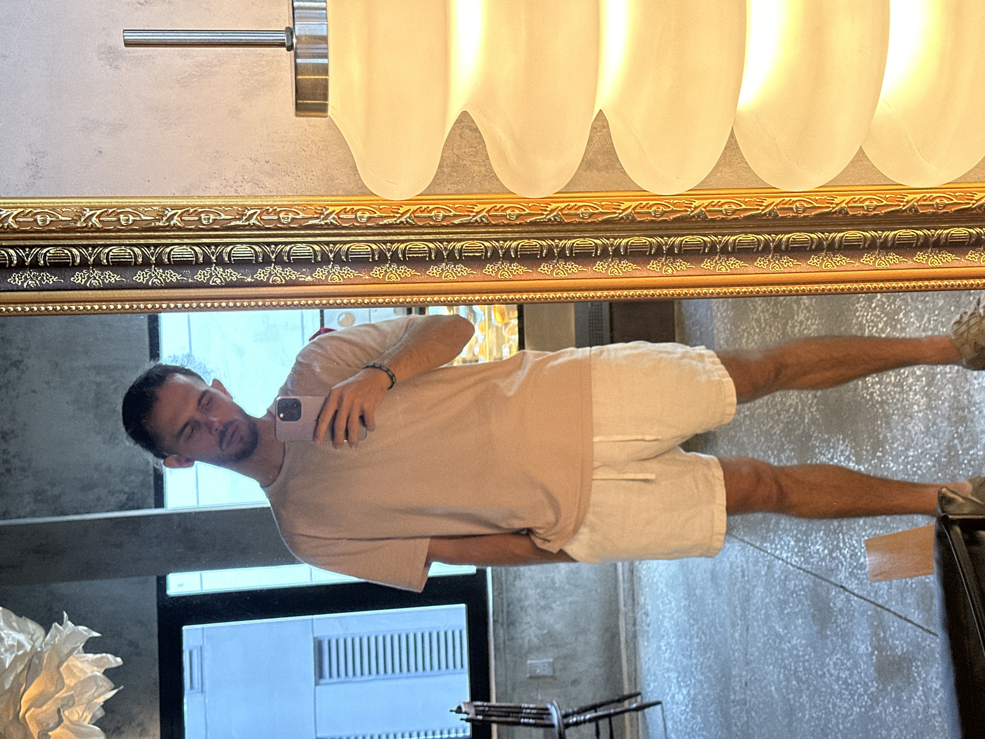 A person is standing in front of a mirror, taking a selfie. The person is wearing a light-colored t-shirt and white shorts. They have their hair pulled back and are looking down at their phone. The mirror has an ornate gold frame. To the right of the mirror is a large, off-white lamp with a ribbed design. In the background, through the mirror, is a window showing a building and what appears to be a display case with food items. The floor is speckled gray. The overall setting appears to be an indoor commercial space, possibly a cafe or restaurant, in Da Nang, Vietnam.