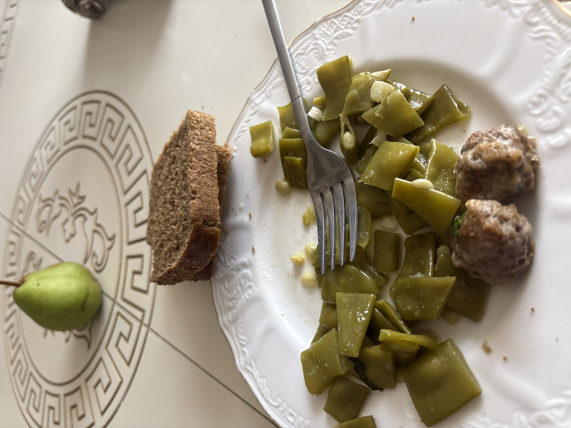 A high-angle, close-up shot shows a white plate with a decorative embossed rim, containing a portion of green beans and two meatballs. A fork rests on the plate, its tines partially submerged in the green beans. Two slices of dark bread are stacked behind the plate. In the background, a light-colored tiled surface features a circular decorative pattern. A green pear is positioned on this surface to the left of the plate. The scene appears to be a casual meal setting.
