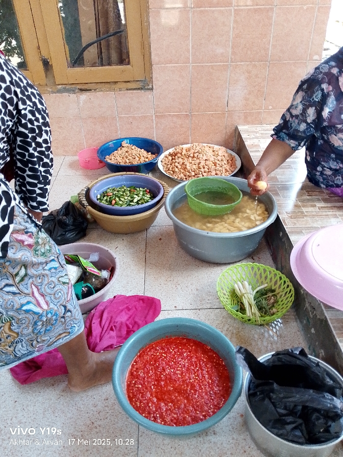The scene is an outdoor patio in Gegesik, Indonesia (latitude: -6.538, longitude: 108.43). Two women, one with dark and one with light skin, are preparing food. One woman is standing, wearing a black and white polka-dot top and a floral patterned skirt, holding a light-colored vegetable, and appears to be washing it. The other woman, wearing a floral patterned top, is out of frame. In front of them are several bowls, including one filled with water, one with chopped green vegetables, one with diced red pepper, and two with small, orange-colored, chopped pieces. There are two plastic baskets, one green and one beige, one containing a large, green vegetable. A pink towel lies on the floor next to a large pot with a black plastic bag in it. The environment is tile, a brown, wooden door, and a window.
