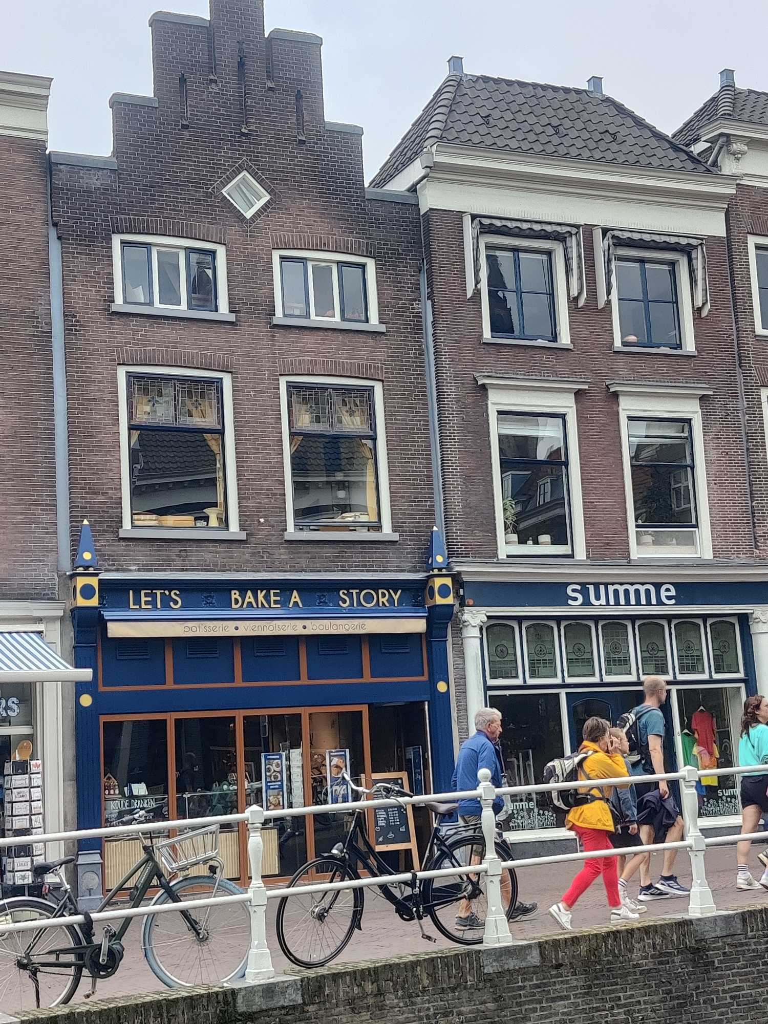 The image shows a street scene in Delft, Netherlands, focusing on the facades of two brick buildings and the activity on a canal-side walkway. The building on the left features a stepped gable and a sign above a blue storefront that reads "LET'S BAKE A STORY" with smaller text below indicating "patisserie • viennoiserie • boulangerie." The building on the right has a more traditional roofline and a similar blue storefront with a sign that says "summe."
Several people are walking along the elevated walkway in front of the shops, separated from the canal by a white railing. From left to right, a man in a blue jacket is walking, followed by a woman in a yellow jacket and red pants, then a man with a backpack, and finally a woman in a turquoise top. Two bicycles are parked on the walkway, one dark-colored and the other olive green. The storefronts display various goods, and reflections in the windows reveal more of the interior and the street beyond. The overall atmosphere suggests a typical day in a European city with shops and pedestrian traffic.