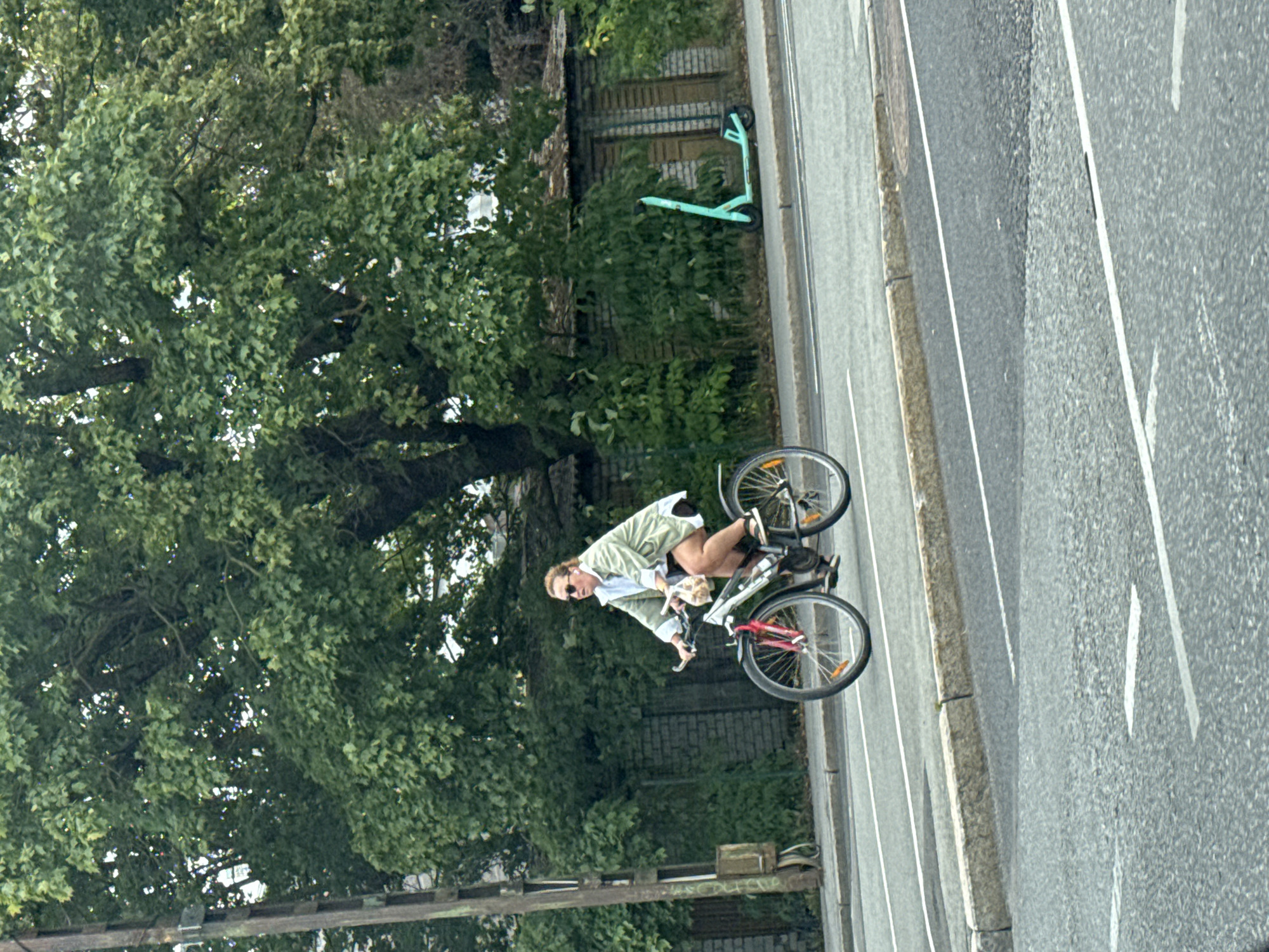 The image shows a woman riding a bicycle on a city street in Tallinn, Estonia. The woman is wearing a light-colored jacket, sunglasses, and headphones. She is carrying a bag of groceries. In the background, there is a turquoise electric scooter parked on the sidewalk. The street is paved and there are lane markings. There are trees and buildings in the background. The sky is cloudy.