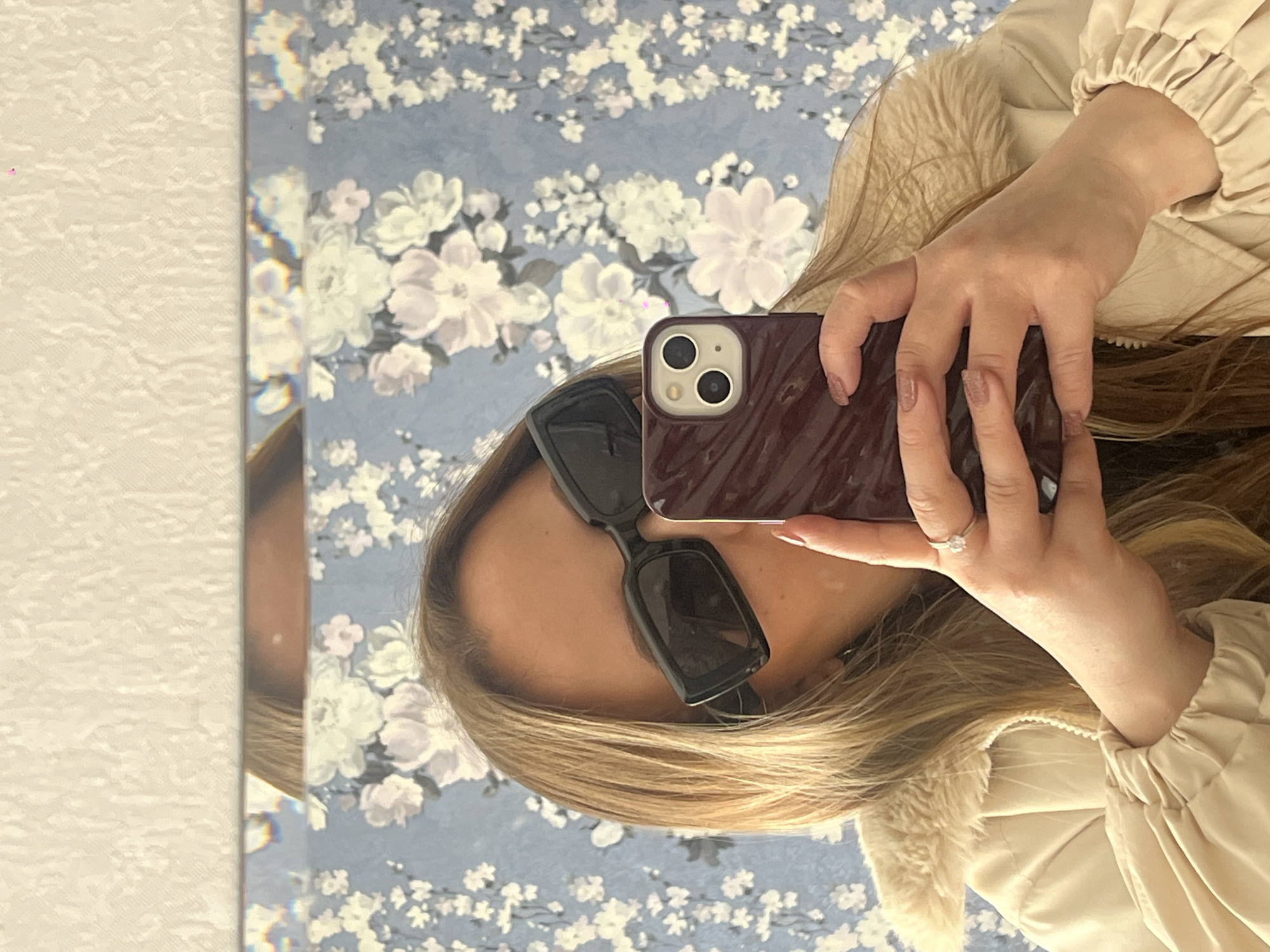 A fair-skinned individual is taking a selfie in front of a mirror with a floral-patterned background. They are wearing sunglasses and a light-colored jacket with a furry lining. The individual is holding a dark-colored smartphone with a dual-lens camera in their left hand. A ring is visible on the ring finger of their left hand. The scene appears to be indoors, with a textured wall visible to the left of the mirror. There is no evidence indicating that this image was taken in Novopokrovskaya, Russia.