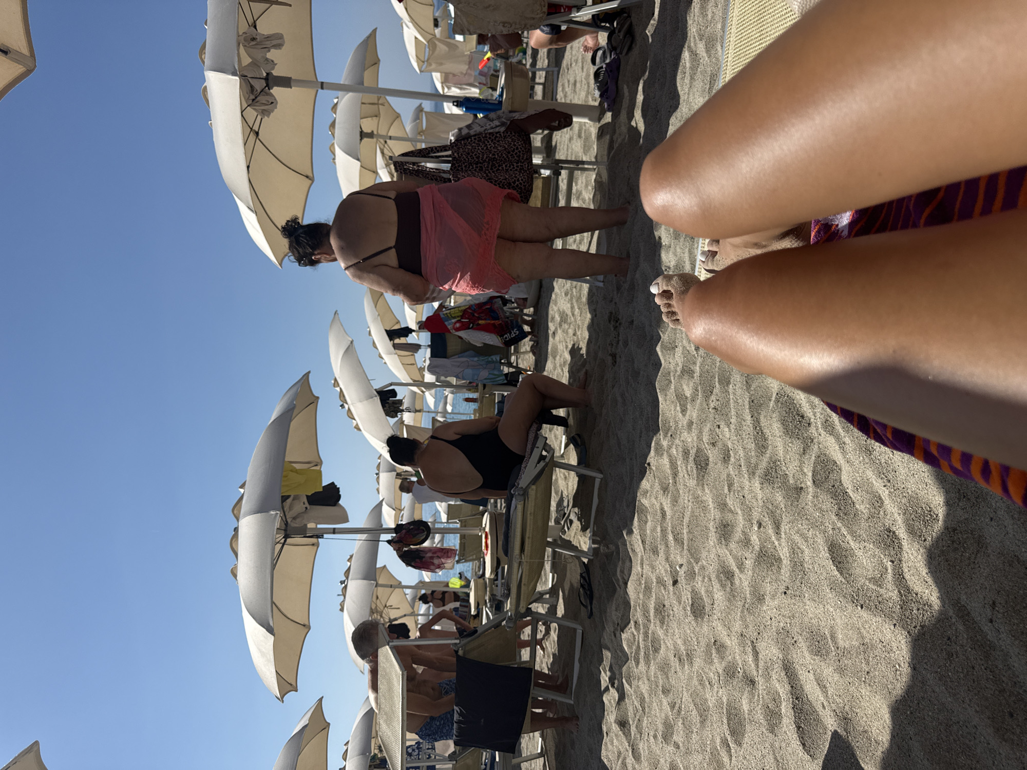 The image depicts a beach scene in Finale Ligure, Italy. The foreground shows a person's legs and feet lying on a beach chair, with sand visible. In the midground, several people are relaxing on the beach, some sitting on lounge chairs and others standing. Beach umbrellas are spread across the sand, providing shade. The background reveals the blue sky and a body of water, likely the Ligurian Sea. The atmosphere is sunny and relaxed, suggesting a typical day at the beach.