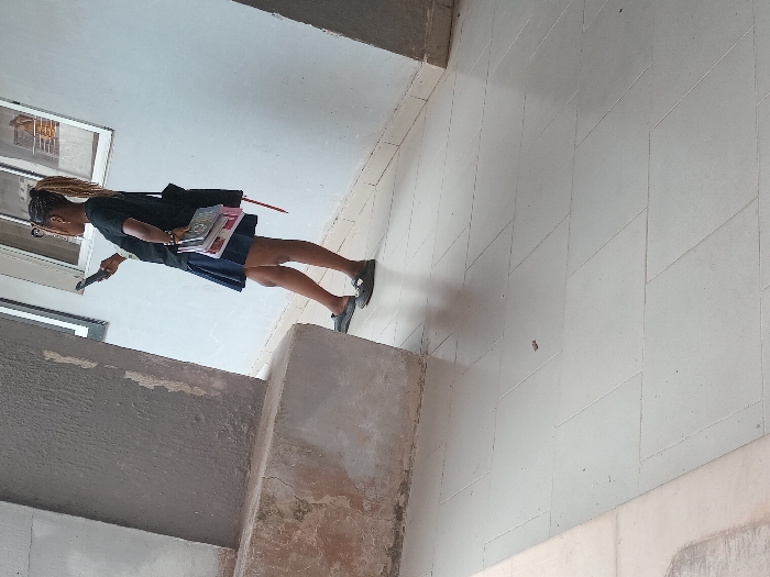 The image captures a female individual standing in a covered walkway of a building in OGBOKO, Nigeria. The subject, observed in right profile, wears a black short-sleeved t-shirt, a dark blue skirt, and dark flat shoes. She has braided hair pulled back and wears eyeglasses. In her left hand, she holds and views a black smartphone. Her right hand carries a stack of approximately four books/notebooks and a dark shoulder bag with a visible red strap. She stands with her weight primarily on her right foot, her left foot slightly forward with the heel raised. The environment consists of a light-colored tiled floor arranged in a diagonal pattern. A light-colored wall with two rectangular, dark-framed windows is visible on the right; one window reflects a metal railing. A thick, textured concrete pillar, light brown-grey in color, occupies the left foreground, showing some minor surface discoloration at its base. The scene is brightly illuminated, suggesting natural light.