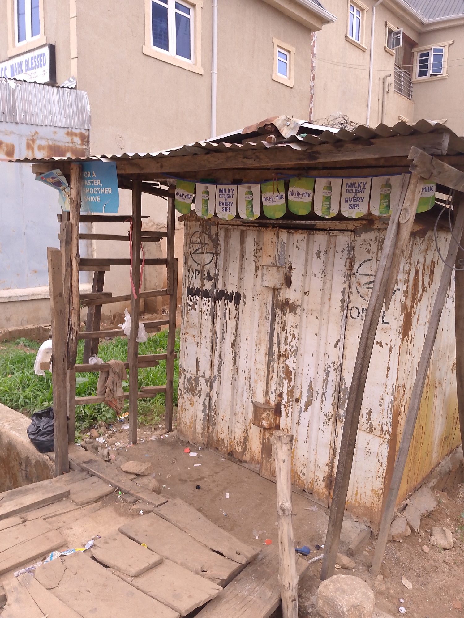 The image depicts a dilapidated kiosk in what appears to be Bukuru, Nigeria, based on the advertising. The structure is made of corrugated metal and wooden supports, with a rusty corrugated metal roof. The front of the kiosk is adorned with banners advertising "Aktiv-Milk" and "Milky Delight," along with a shaving product ad. The door of the kiosk has peeling paint and the word "OPEN" spray-painted on it. The kiosk sits on a wooden platform with missing and broken planks. The surrounding area is littered with debris and trash. In the background, a modern building with windows is visible. A handmade wooden ladder leans against the left side of the kiosk.