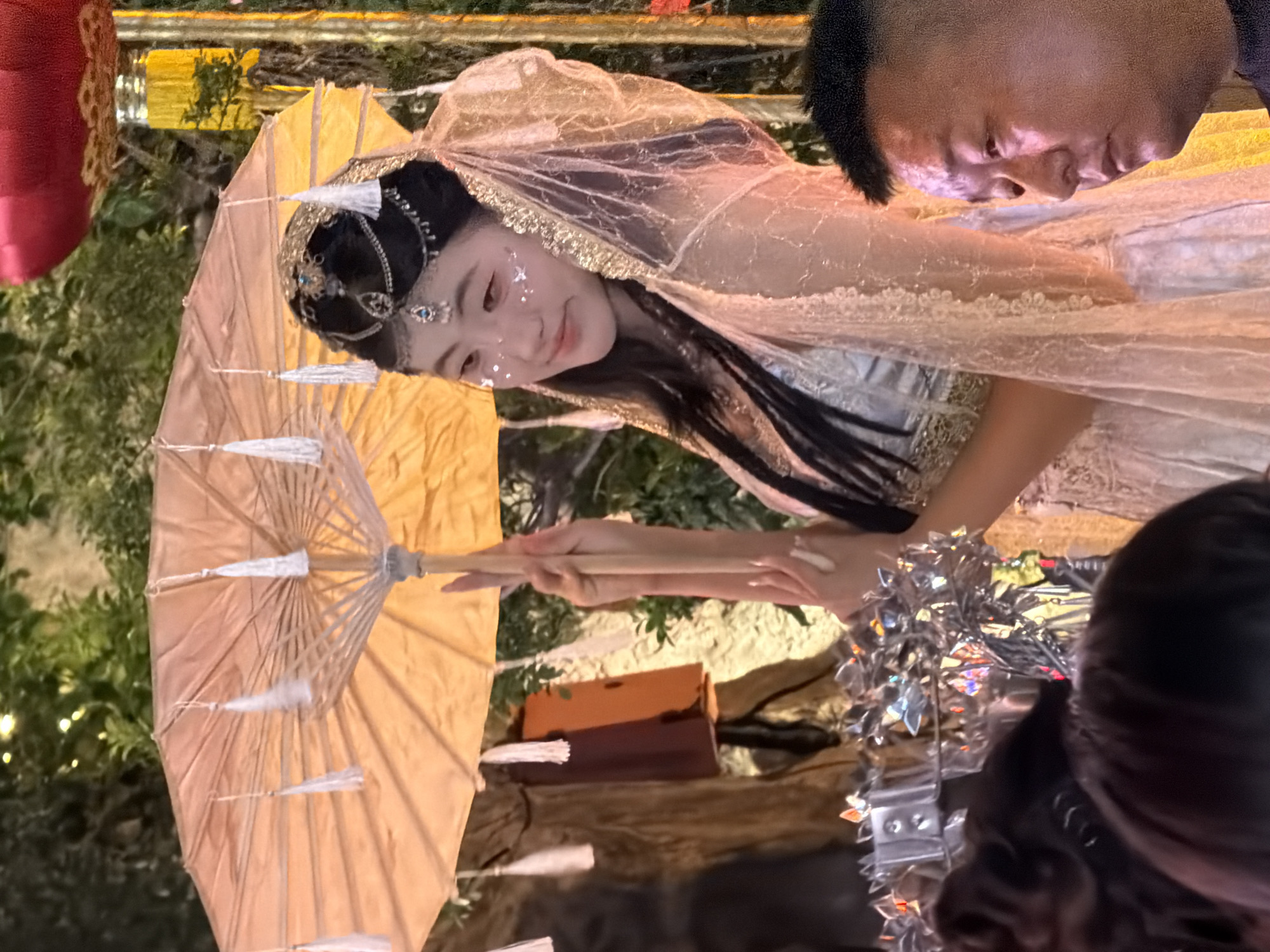 In the Yuzhong District of China, a young woman adorned in traditional attire, complete with a jeweled headdress and shimmering makeup, holds an ornate parasol. She is dressed in a light-colored, intricately embroidered gown with a delicate veil draped over her shoulders. The parasol, an elegant pale yellow, features white tassels hanging from its spokes. Behind her, lush green foliage and decorative red lanterns suggest a festive outdoor setting, possibly a cultural event or celebration. In the foreground, the side profile of a man is visible, his face illuminated by warm lighting. The scene is rich with cultural detail and a sense of occasion.