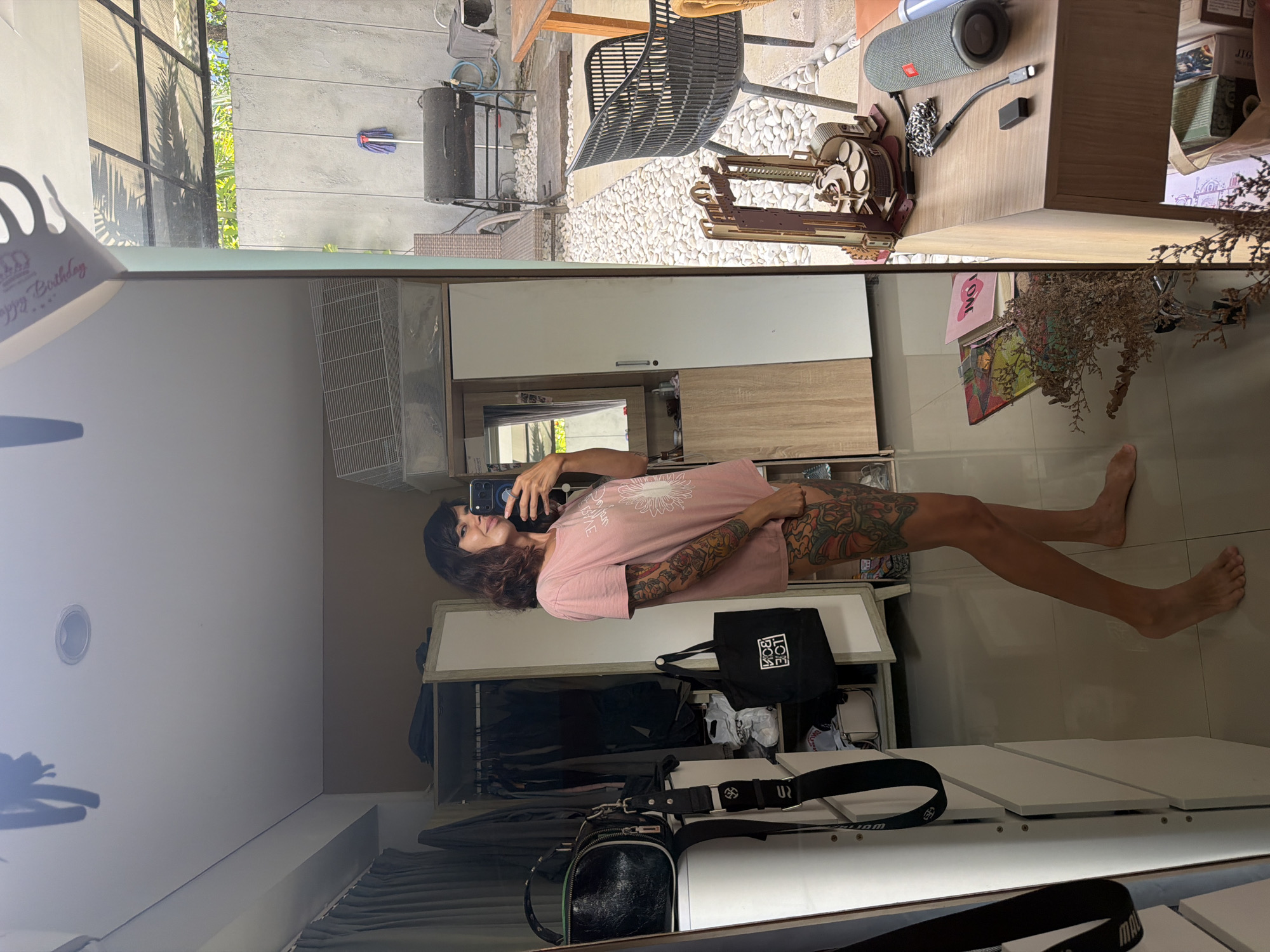 The image captures a full-body mirror selfie taken indoors, reflecting an outdoor scene beyond. The subject is an adult with dark, wavy hair, wearing a light pink t-shirt with a white circular design, possibly a stylized flower or sun. They have extensive, colorful tattoos covering their arms and legs, and are holding a smartphone to take the picture. Their bare feet are visible on a light-colored tiled floor.
The indoor reflection reveals a room with modern white and wood-toned cabinetry. A black tote bag with the white text "LOVE BOX" is visible on a shelf, and a black cross-body bag with a strap partially showing "WORLDWIDE" lies on a lower surface. A dried floral arrangement and several framed pictures or art pieces are displayed on a shelf. A light fixture is embedded in the ceiling.
Beyond the room, a bright outdoor area is reflected, suggesting a large window or glass door. This space features white stone aggregate landscaping, a black metal chair, and a woven rattan chair. A wooden table holds a grey JBL Flip speaker, a charging cable, and an intricate wooden mechanical model, resembling a historical gun or crossbow. Lush green tropical foliage and trees are visible, along with a concrete wall where a blue mop hangs, and a large black speaker or container is placed.
The scene is brightly lit, indicating daytime. A partially visible "Happy Birthday" crown is in the immediate foreground. The setting appears to be a residence or guesthouse in Kuta Selatan, Indonesia.