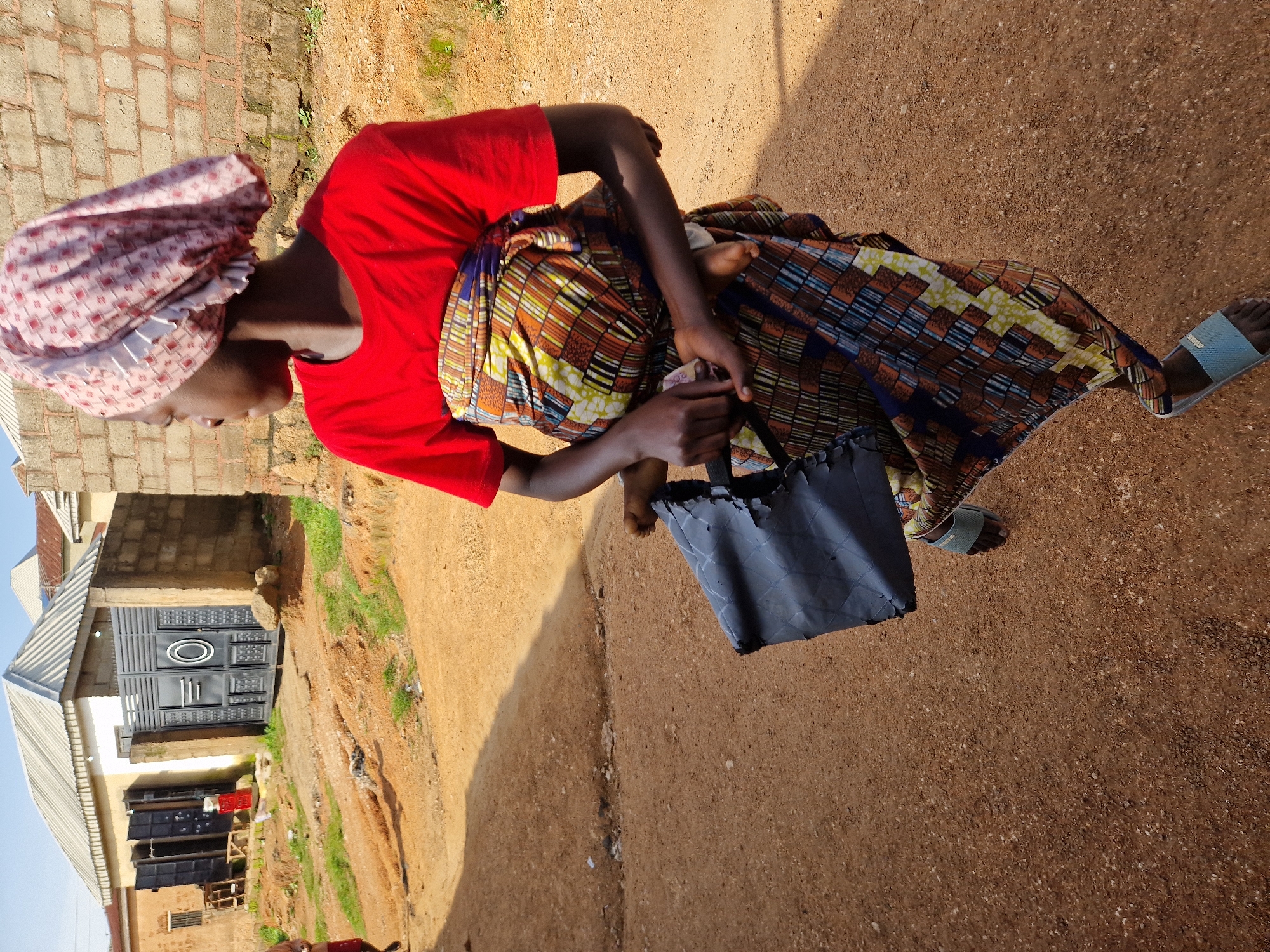 A young person wearing a red t-shirt and a pink headscarf is standing outdoors in what appears to be a street or pathway in Jos, Nigeria. They are carrying a baby wrapped in a colorful fabric around their waist and holding a black plastic bag. The background shows a building with a metal gate, a brick wall, and some vegetation. The ground is unpaved and appears to be dirt. The lighting suggests it is daytime.