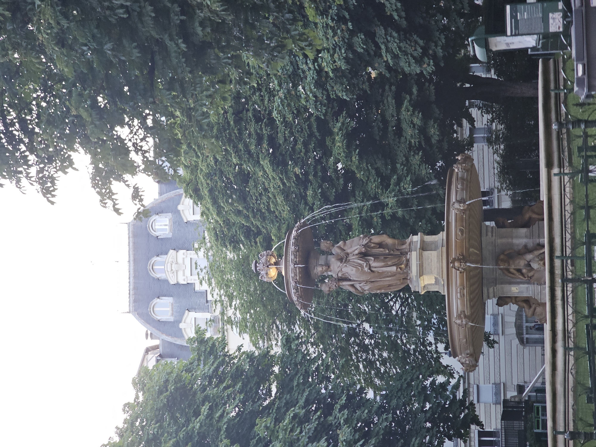 The image shows the Fontaine Saint-Sulpice, a monumental fountain located in the Place Saint-Sulpice in the 6th arrondissement of Paris, France. The fountain is made of bronze and stone, with statues of four prominent French clergymen: Bossuet, Fénelon, Fléchier, and Massillon. Water cascades from the upper basin into the lower one. Trees surround the fountain, and a building is visible in the background. The image is taken during daylight hours, although the light seems diffused. The orientation of the image is rotated.