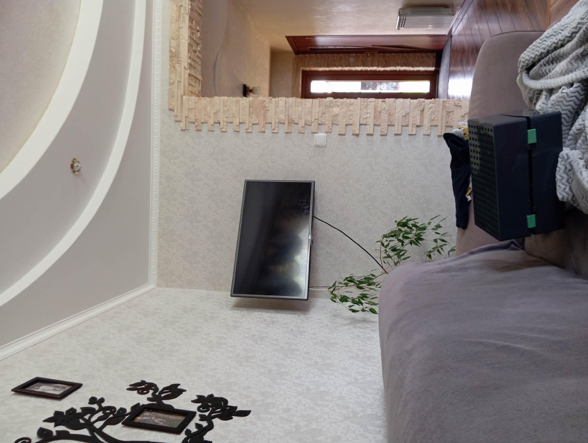 The image depicts an interior scene, likely a living room, with elements of a hallway visible in the background. In the foreground, a portion of a sofa is covered with a light purple blanket. To the right of the sofa, a dark grey box with green accents and a black patterned lid rests on the furniture. A light-colored, textured throw is draped over the sofa in the right foreground.
The wall behind the sofa is covered with light-colored wallpaper with a subtle floral pattern. A flat-screen television is mounted on the wall, displaying a dark, reflective surface. A small potted plant with green leaves is positioned below and to the left of the television. A black wire extends from the television down the wall.
To the left of the television, a black decorative tree silhouette with attached picture frames is affixed to the wall. Two framed pictures are visible within the silhouette. The ceiling is a two-tiered design with a curved, recessed central section, illuminated by a single light fixture.
A stone-tiled column or archway is present on the right side of the image, separating the living area from a hallway. In the hallway, a wooden door with glass panes is visible, suggesting an entrance or another room. Further down the hallway, a second wooden door is partially visible, and a white radiator is mounted on the wall. The floor in the hallway appears to be polished wood. The environment suggests a residential setting in Hlynytsi, Ukraine.
