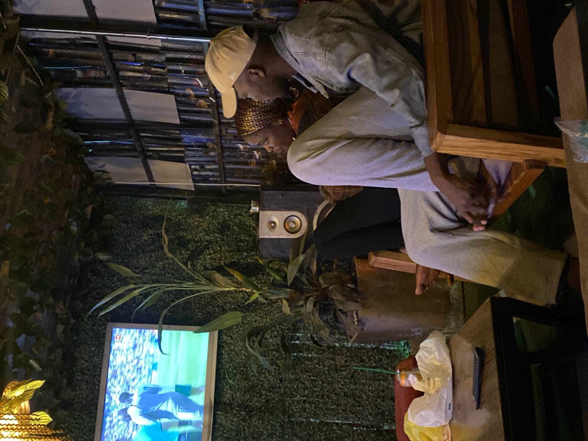 In Gura Top, Nigeria, a couple watches a soccer match on a television screen mounted on a wall covered in artificial greenery. They are seated on wooden chairs in what appears to be an outdoor or semi-outdoor space, possibly a cafe or lounge area. The male, wearing a light-colored baseball cap and a denim jacket, sits to the right, his legs crossed. The female, to his left, is partially obscured but wears a headscarf with a repeating pattern. Between them and the television, a potted plant with long, green leaves stands in a rustic pot. A speaker is visible to the right of the plant. In the foreground, a dark wooden table holds a remote control, a can, and some white paper packaging. The background features a wall composed of vertical metal elements, some covered with white material, and further artificial greenery draped from the ceiling.