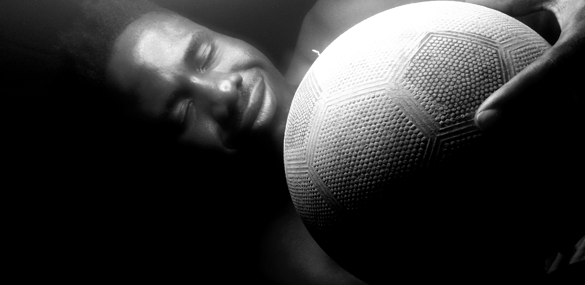 The black and white image depicts a man holding a white soccer ball. He is dark-skinned and appears to be resting, with his eyes closed. The ball is close to his face, and a hand is visible holding it. The setting is dark, with a stark contrast between the illuminated areas and the shadows. No specific location is discernible, and it is not clear this takes place in Narayi, Nigeria.