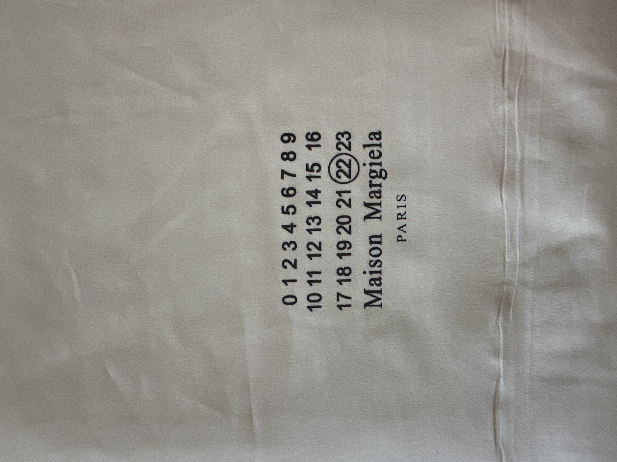 The image displays a close-up, frontal view of a beige fabric material with black text printed on it. The text features a numerical sequence arranged in rows, followed by the words "Maison Margiela" and "PARIS". The numbers are arranged as follows: "0 1 2 3 4 5 6 7 8 9" on the first line, "10 11 12 13 14 15 16" on the second line, and "17 18 19 20 21 22 23" on the third line. Notably, the number "22" in the third row is enclosed in a circle. The fabric exhibits soft folds and creases, creating subtle shadows and highlights across its surface. The lighting appears to be diffuse, illuminating the fabric evenly. The background is entirely the beige fabric material, with no other elements or environmental context visible. There are no people, animals, or other objects present in the image. The image has not been geolocated to Bečići, Montenegro.