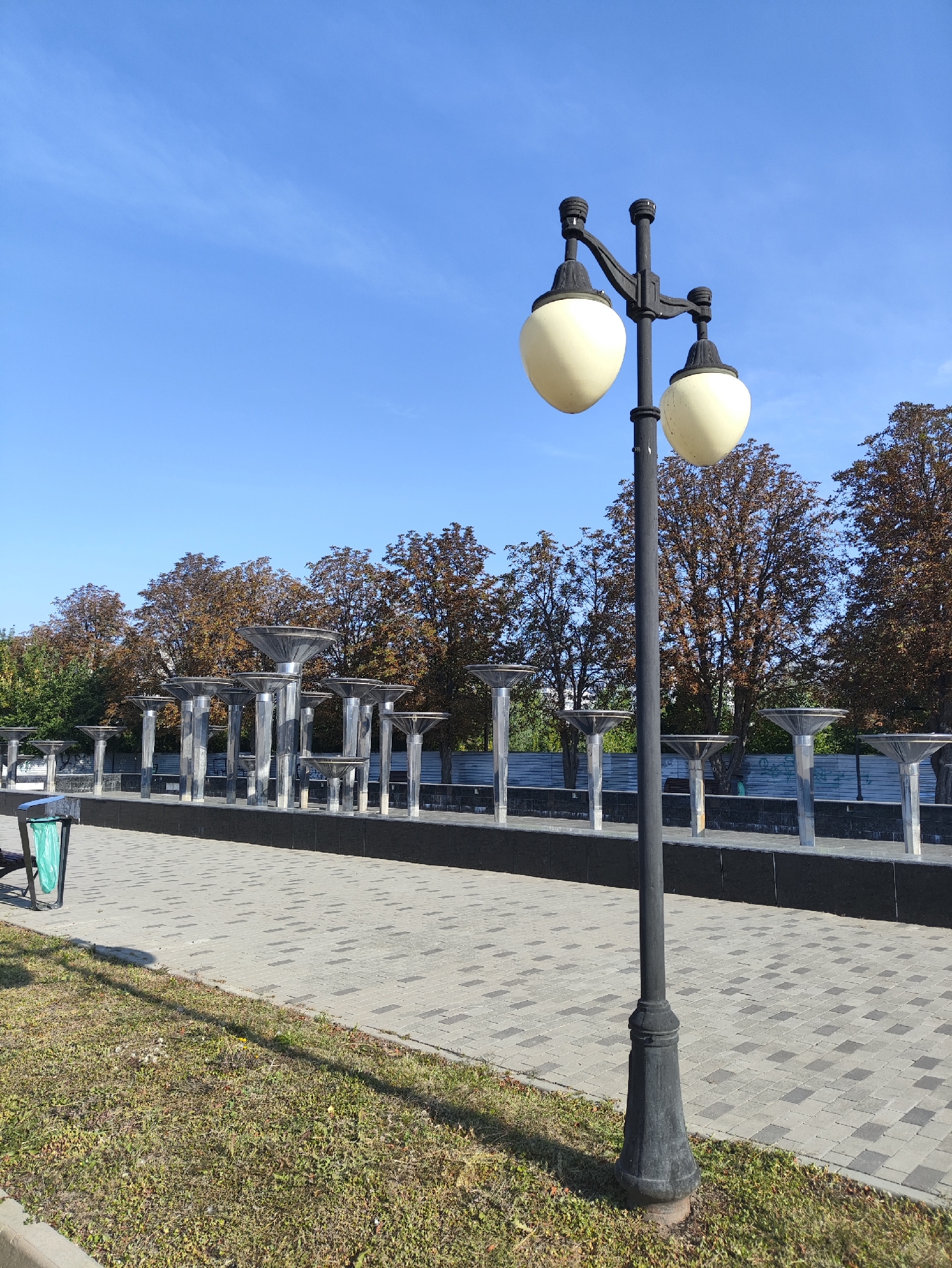 The image depicts a park scene on a clear day with a bright blue sky. In the foreground, a black street lamp with two cream-colored globe lights stands prominently. The lamp is positioned on a paved walkway made of gray interlocking bricks, which extends into the midground. To the left of the walkway, a patch of green grass with some dry patches and small yellow flowers is visible.
Behind the walkway and lamp, a long, modern fountain structure is present. It consists of a series of sleek, metallic cylindrical columns topped with wide, cone-shaped basins. The fountain appears to be dry. Behind the fountain, a line of trees with autumn foliage in shades of brown and orange can be seen, with some green trees interspersed. A gray metal fence runs behind the fountain and trees. In the far left of the image, a green trash can is visible next to the walkway. The overall impression is that of a well-maintained public space. The location is indicated to be in the city of Kharkiv, Ukraine.