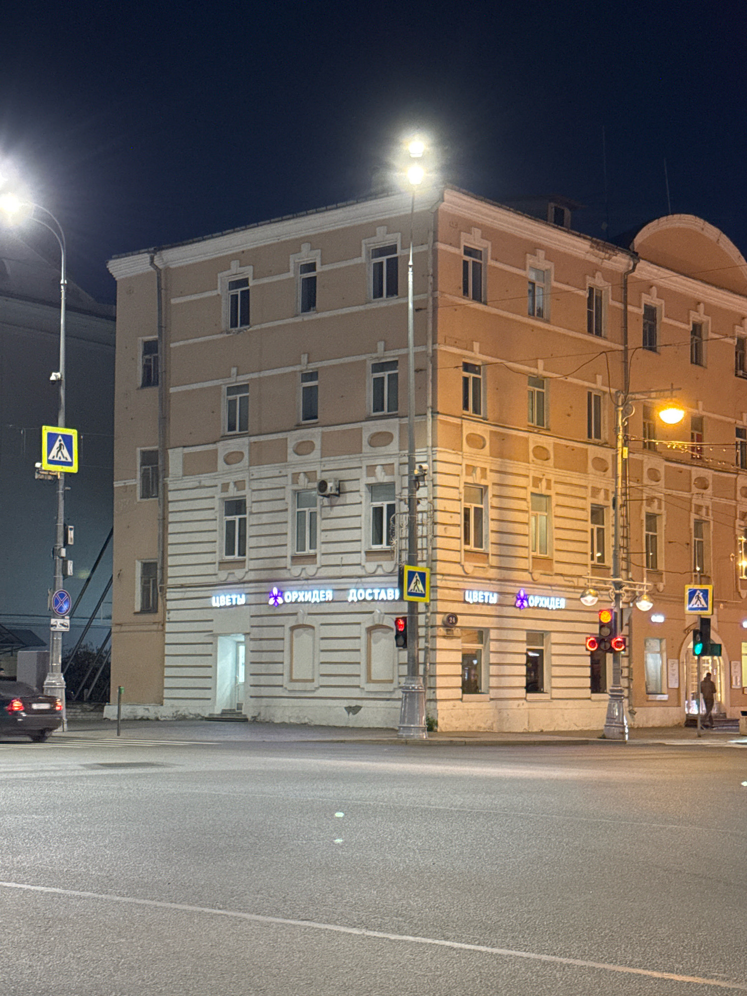 The image shows a nighttime view of a street corner in Tver, Russia. The foreground is dominated by a wide, empty asphalt road with faint white lane markings. In the middle ground, a multi-story building with a light beige facade stands prominently. The building features numerous windows and decorative elements. On the ground floor of the building, there are signs with Russian text and a purple orchid logo, indicating a flower shop. Several streetlights illuminate the scene, casting a warm glow. Traffic lights are visible, one showing red and others showing green, suggesting an intersection. A pedestrian crossing sign is mounted on a pole. A dark-colored car is partially visible on the left side of the frame, and a person is seen walking on the sidewalk on the right side. The sky above is dark, indicating nighttime.