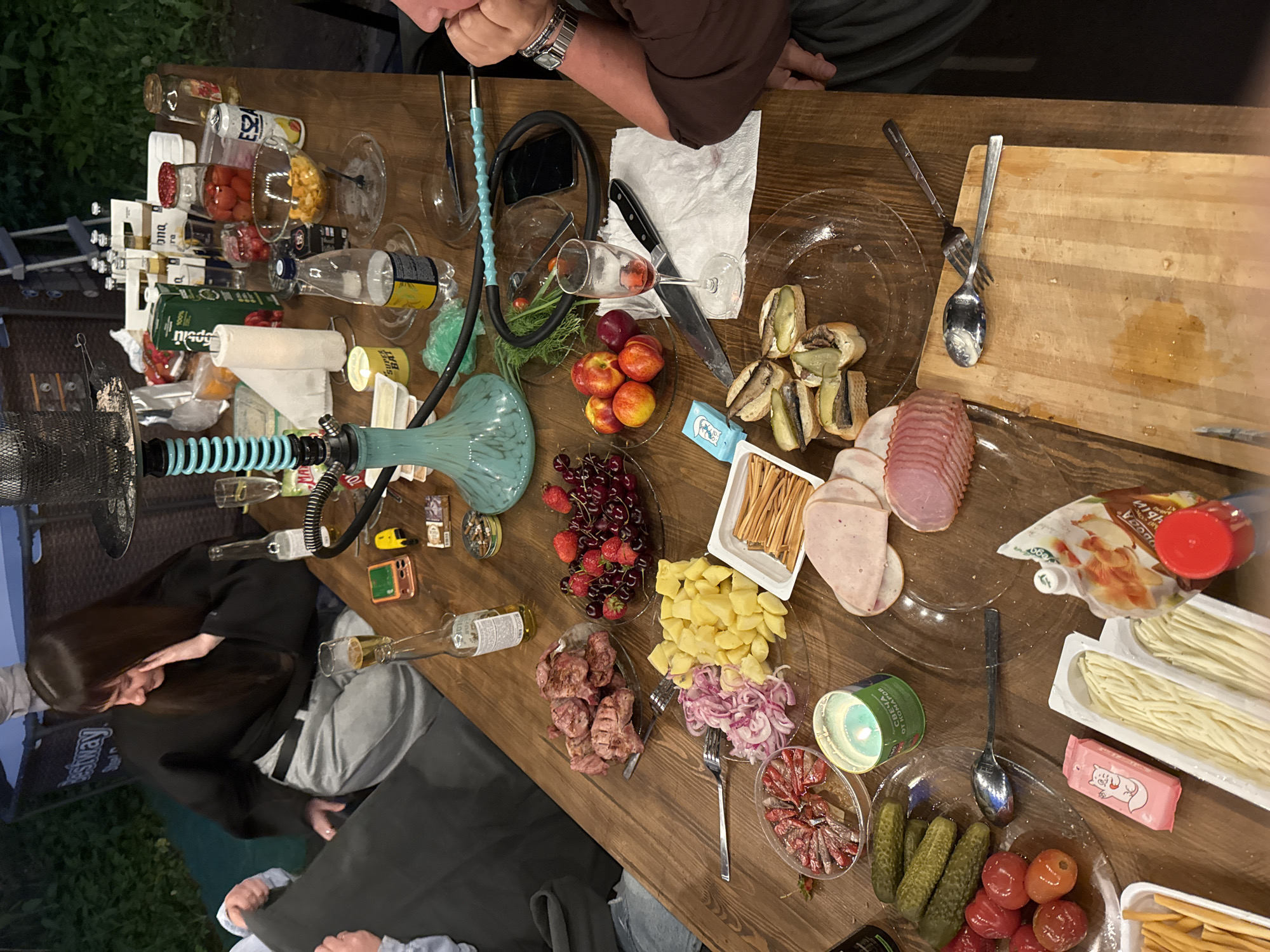 The image depicts an outdoor evening gathering around a large wooden table in Bakhtiyary, Russia. The scene is illuminated by artificial light, suggesting dusk or nighttime. **People:** Four individuals are partially visible. On the left, a woman with long dark hair, wearing a black long-sleeved top and light grey pants, is seated, looking down with her left hand touching her face. Next to her, a person's arm with a light blue sleeve is visible. On the right, a person's arm and shoulder are visible; they are wearing a dark brown long-sleeved shirt and a silver wristwatch, with their hand near their chin. Another person's arm is visible at the top left, reaching into the frame. **Objects on the Table (from left to right):** The dark wooden table is densely covered with food, drinks, and various items. * **Hookah:** A prominent light blue and black hookah with a black hose and light blue mouthpiece is centrally located on the table. * **Beverages:** Multiple beer bottles, including "Corona Extra," are in a white cardboard carrier. A green carton of juice (marked "100%"). A can of "ESZA." Several plastic water bottles. A glass of amber liquid (likely beer) is next to the woman on the left. A wine glass containing red residue (likely red wine) is positioned near the person on the right. * **Food:** * Plates contain grilled meat (likely pork shashlik), cubed potatoes, sliced red onions, fresh strawberries and cherries, and several nectarines/plums. * Small open-faced sandwiches with sprats or sardines and pickled cucumber slices are on a plate. * A transparent container holds thinly sliced red peppers and possibly pickled fish. * A bowl contains light yellow crunchy snacks, possibly chips or croutons. * A plate holds a quantity of small pickled cucumbers. * Another plate contains cherry tomatoes. * A rectangular plastic container holds long, thin, light-colored cheese or snack sticks. * A platter features various sliced cold cuts (e.g., ham, salami). * A white squeeze pouch of mayonnaise with mushrooms (Майонез с грибами) and a red-capped plastic bottle (likely for condiments) are present. * A pink packet with a squirrel mascot ("Белочка" candy) and a small blue box labeled "ОХОТНИЧЬИ" are also on the table. * **Tableware and Accessories:** * Numerous clear glass plates, some empty, some laden with food. * Forks, spoons, and knives are scattered across the table, including a large chef's knife next to the