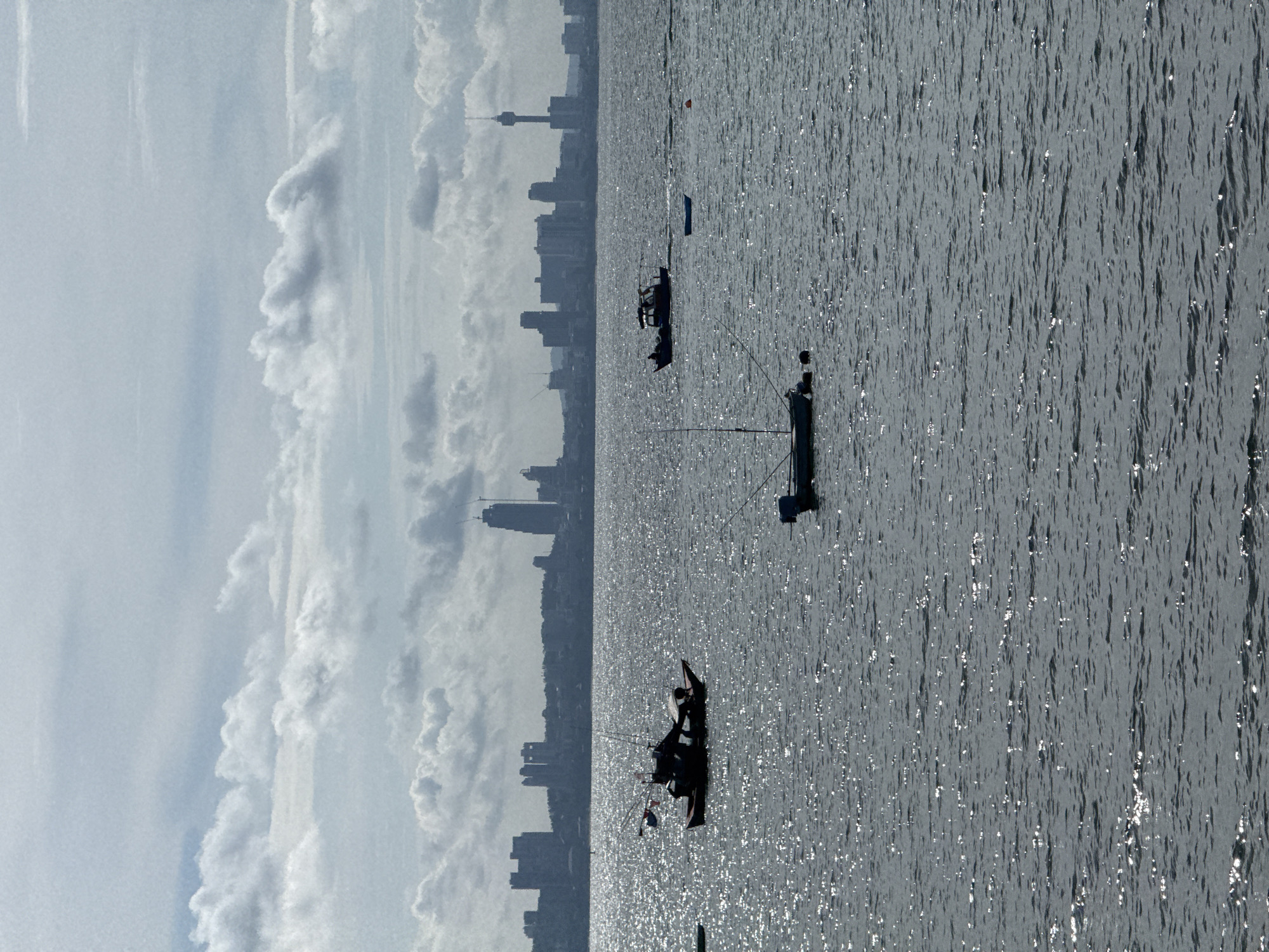The image depicts a seascape off the coast of Pattaya City, Thailand. Three fishing boats are visible on the water, with silhouettes of individuals aboard. The city's skyline, including a prominent tower, is discernible in the background, obscured by clouds and atmospheric haze. The water surface glimmers with reflected sunlight.