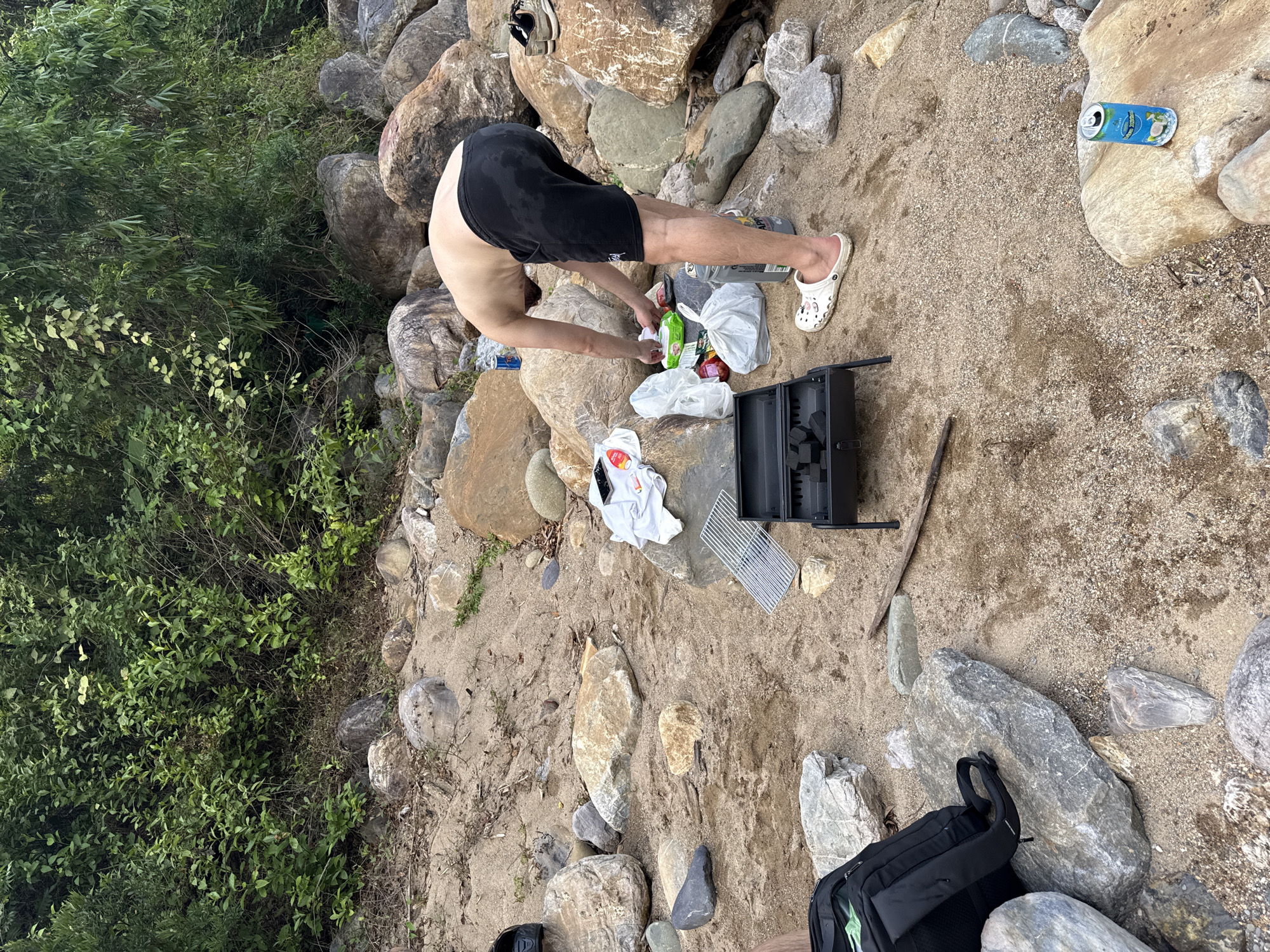 A shirtless man in black shorts and white Crocs is bent over, reaching into a plastic bag on a rocky, sandy area. There is a small black portable grill and a backpack nearby. A blue drink can sits on a rock in the background. Green foliage is visible along the left edge of the image. The location is likely in Đà Nẵng, Vietnam.