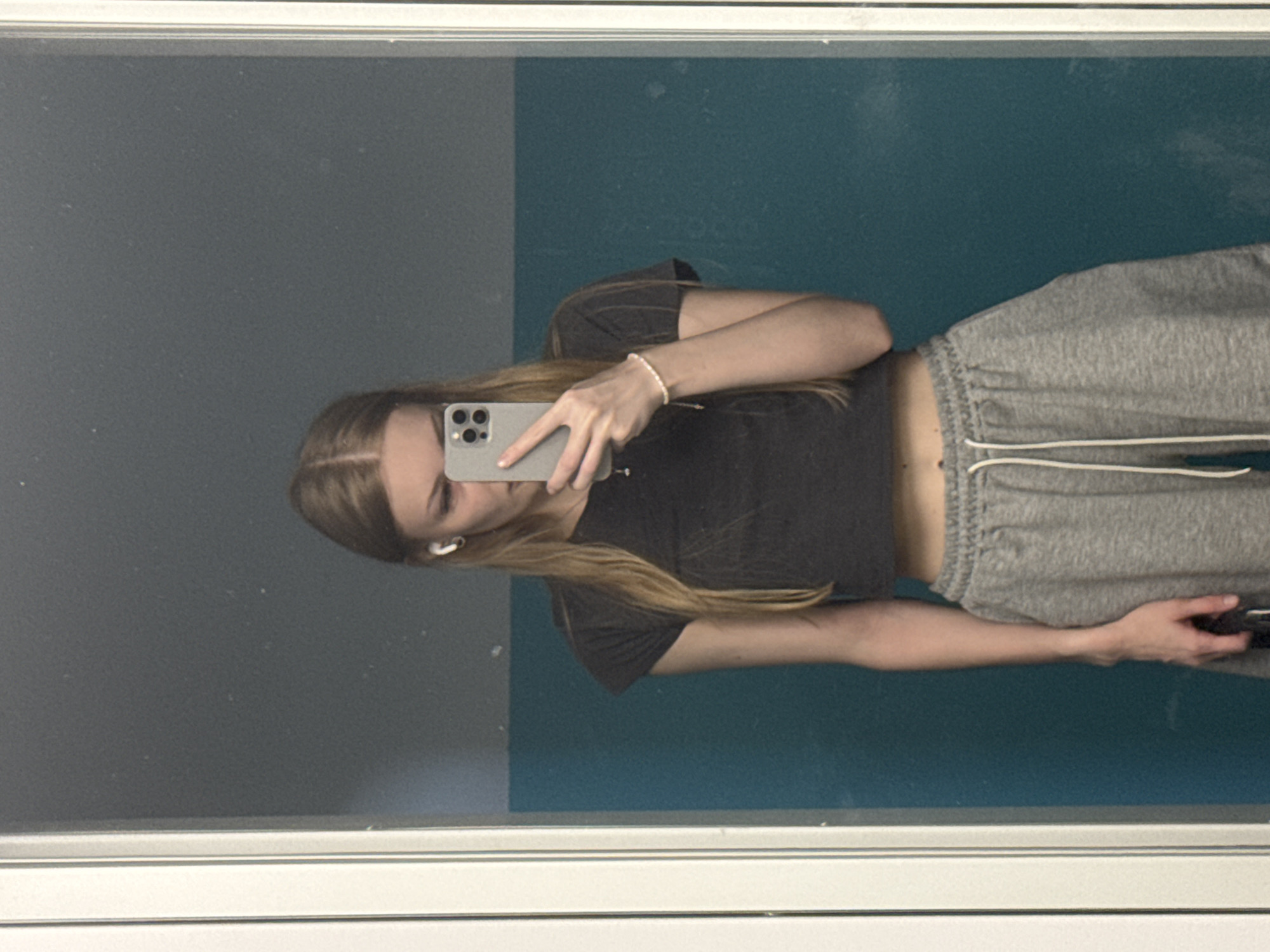 A young woman is taking a selfie in a mirror. She is wearing a dark grey cropped t-shirt and grey sweatpants. She has blonde hair and is wearing white earbuds and a thin bracelet. The background appears to be a hallway with teal and grey walls. The mirror is framed by white molding. It is not possible to determine the location in the city of Perm, Russia from the image.