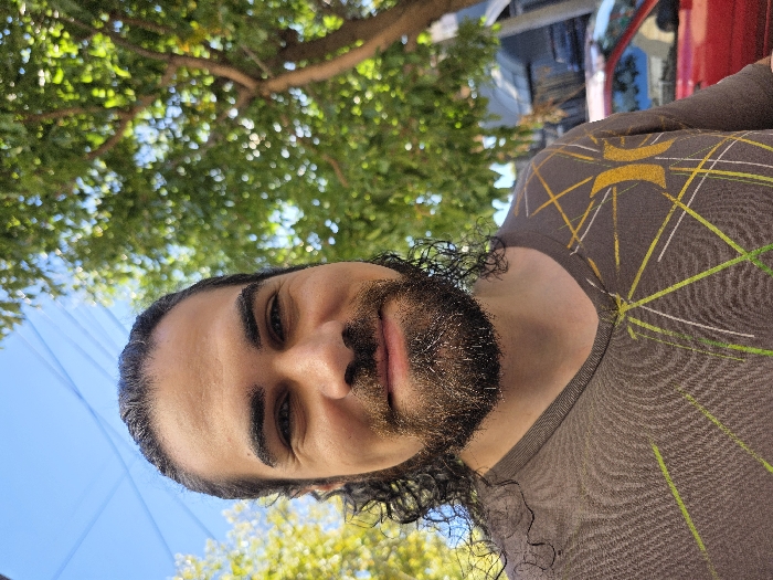 The image displays an outdoor, brightly lit, close-up shot of a single male subject in Villa San Andrés, Argentina. The subject, a man with dark curly hair pulled back, a full dark beard, and a mustache, is centered and smiling directly at the camera. He is wearing a short-sleeved, brown V-neck t-shirt patterned with intersecting thin lines in yellow, white, and light green, and a faded yellow-orange Hurley logo on the right chest area. The background is out of focus. The upper left portion shows a clear blue sky with two thin, dark power lines crossing diagonally. Green foliage from trees is visible to the subject's left and prominently on the right side of the background. A red vehicle is partially discernible in the blurred far background on the right, positioned near a dark, indistinct building or structure. The scene is illuminated by natural daylight.
