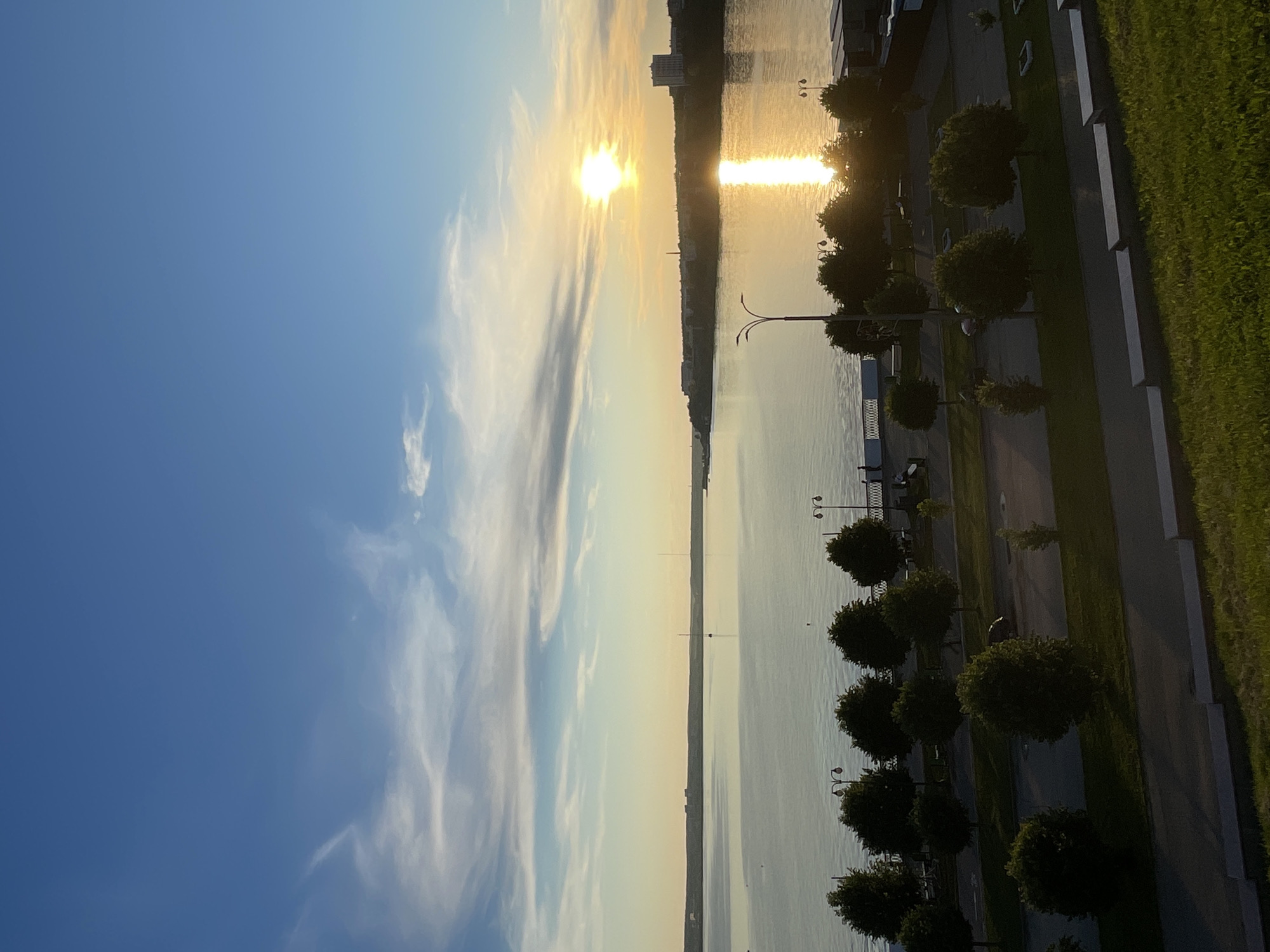 The image depicts a sunset over a wide body of water, likely a river or lake, with a city skyline visible on the distant horizon. The sun is a bright, intense orb low in the sky, casting a golden reflection on the water. Thin, wispy clouds streak across a clear blue sky. In the foreground, there is a tree-lined promenade or park area adjacent to the water. Several streetlights are visible along the promenade, and a few people can be seen walking. The ground in the immediate foreground is covered in green grass. The overall atmosphere is serene and tranquil, characteristic of a late summer or early autumn evening.
