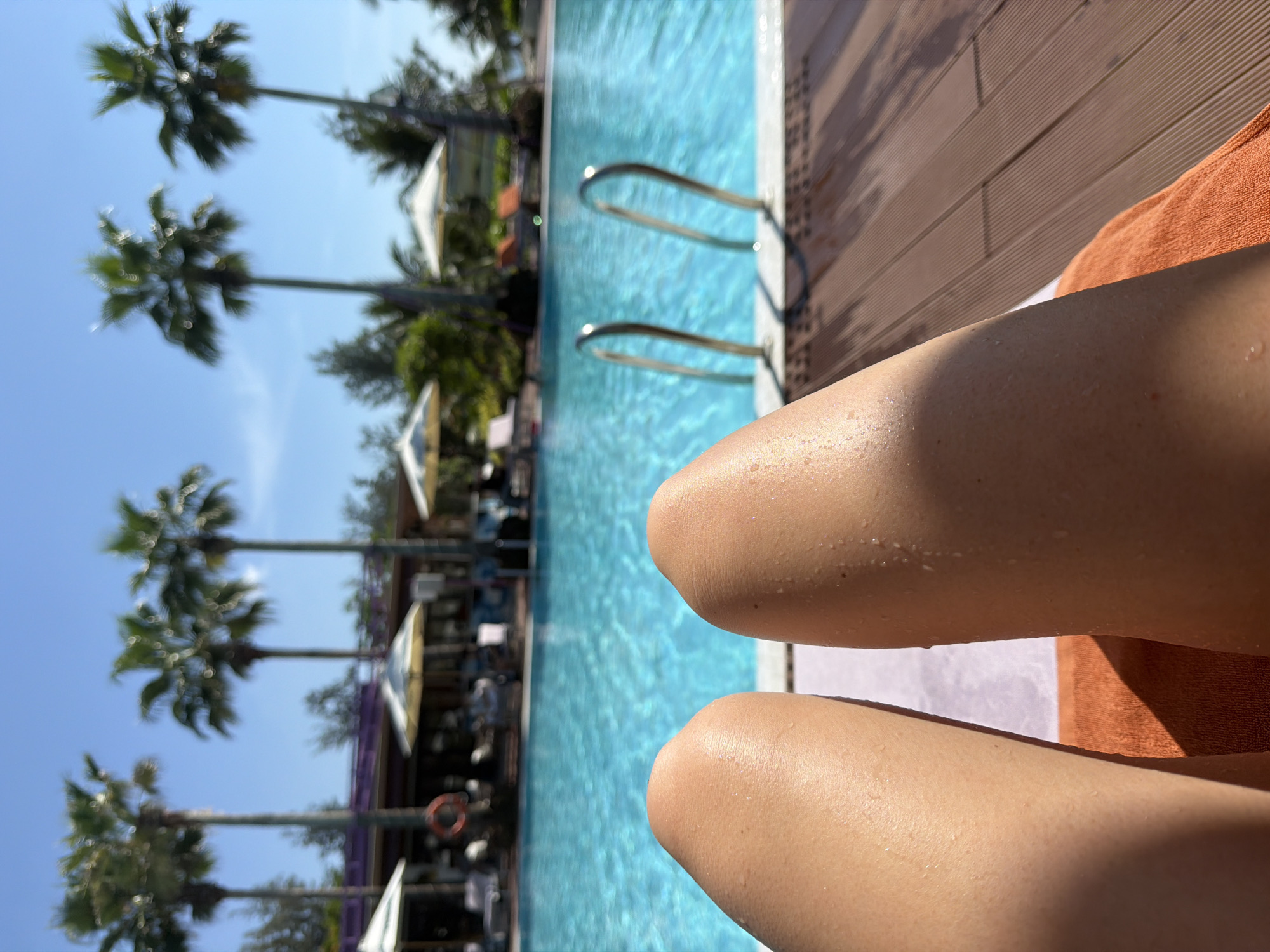 This image shows a close-up view of a person's legs and thighs, positioned in the foreground. The skin appears sun-kissed and has water droplets, suggesting a recent dip in a pool or being in a humid environment. The legs are resting on an orange towel, which is on a wooden deck.
In the background, a clear blue swimming pool is visible, with metal ladders leading into it. Beyond the pool, there are several palm trees casting shadows, and in the distance, there are lounge chairs and umbrellas, indicating a resort or hotel setting. The sky is bright blue and clear, suggesting a sunny day.
The location is identified as a city in Dong Ninh Hoa Ward, Vietnam.