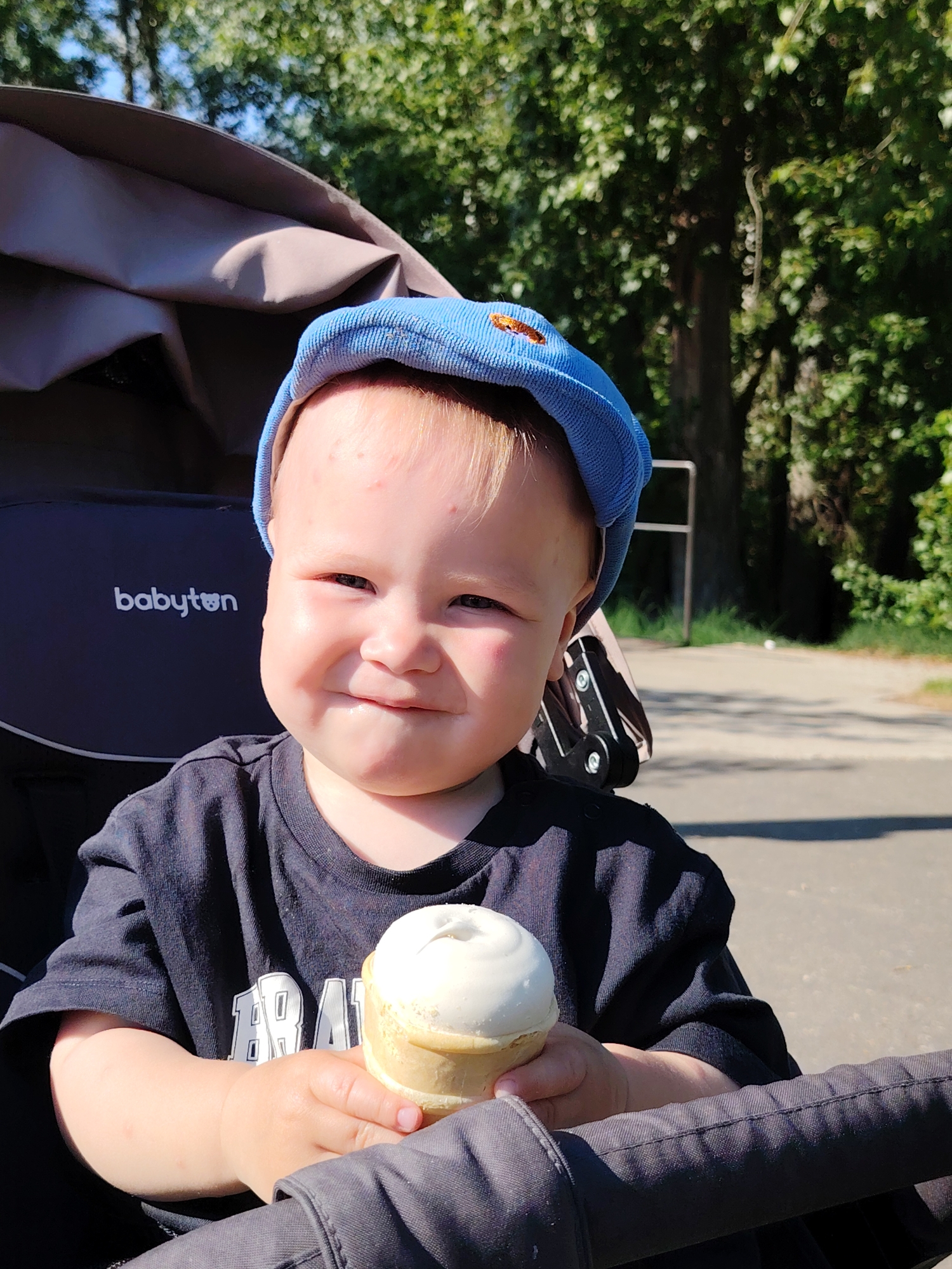 A young child, wearing a blue cap and a black t-shirt, sits in a stroller and holds an ice cream cone. The stroller has a dark gray canopy and the word "babyton" is visible on the side. Trees and a paved walkway are visible in the background. The location is identified as Chaikovsky, Russia.