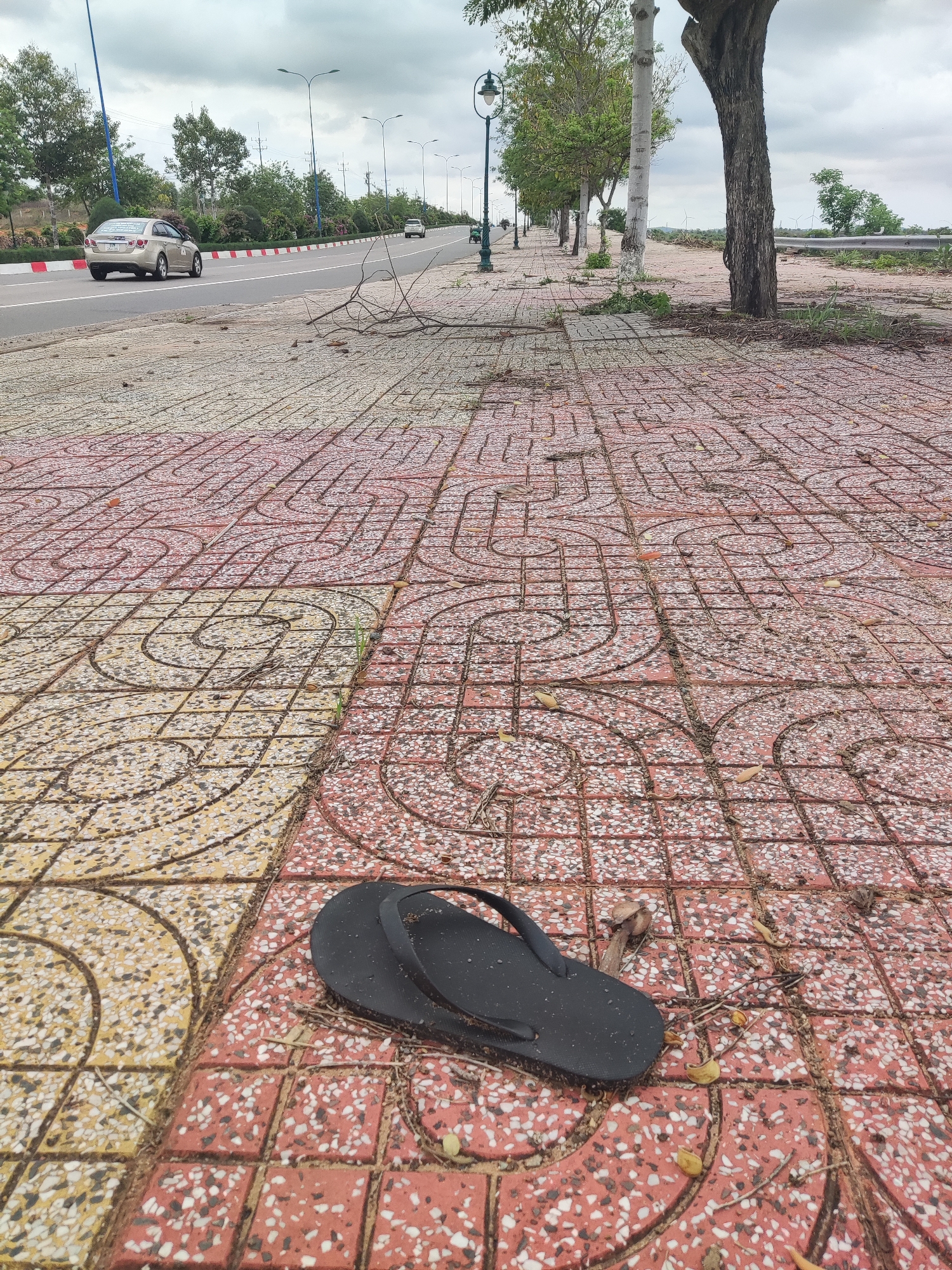 An outdoor, eye-level shot captures a paved sidewalk and a parallel road under a cloudy sky. In the immediate foreground, a single black flip-flop lies on a sidewalk composed of red and yellow patterned tiles, scattered with small leaves and debris.
The sidewalk extends into the midground, lined with trees of varying sizes and black streetlights with lantern-style tops. The road, separated from the sidewalk by a white curb with red and white striped markings, features two lanes of traffic. A light-colored sedan is visible driving away in the left lane, and another white car is further down the road in the distance. Text visible on the rear window of the silver sedan reads "TAXI KHANH HOA," "ANH TUYEN," and two phone numbers: "0913 462 763" and "0903 520 892."
Beyond the trees and road on the right, a metal guardrail runs along the edge of what appears to be a lower embankment. In the far distance, several wind turbines are visible against the overcast sky. The overall scene depicts a roadside area, possibly in a semi-urban or rural setting, with signs of human activity and infrastructure. The time of day appears to be daytime, under heavy cloud cover. No people are visible.