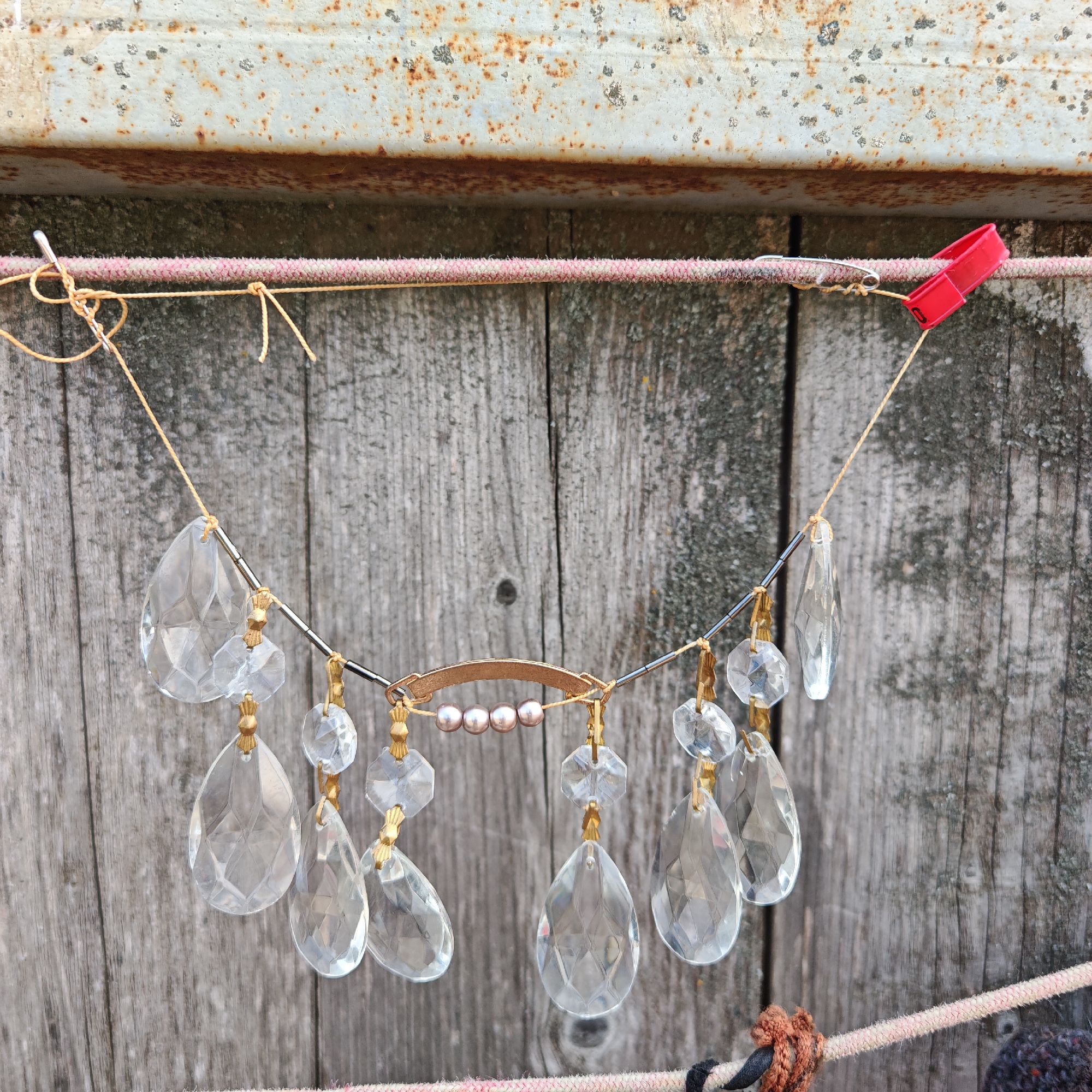 The image shows a decoration hanging on a rope in front of a weathered wooden surface. The decoration consists of a metal rod strung with crystal pendants, glass beads, and a gold-colored curved metal piece holding four pink pearls. The pendants are teardrop and octagon-shaped. The decoration is suspended by a thin, gold-colored string attached to a rope, which is secured on either side by a safety pin and a red plastic clothespin. Above the wooden surface, there's a rusty metal structure. The background consists of weathered wooden planks. The location appears to be outdoors, possibly a backyard or shed. No people are visible in the image. The setting is in the city of Unknown, Unknown.