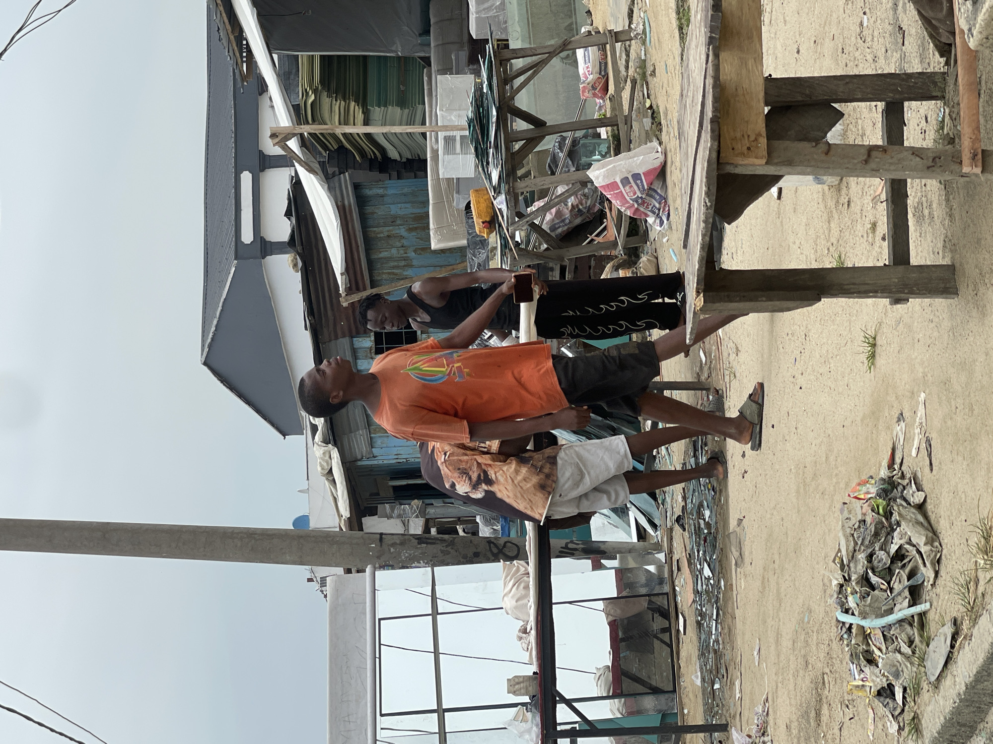 This outdoor image, likely taken in a commercial or market area in Ado, Nigeria, depicts a daytime scene under an overcast sky with dusty ground.
Three men are visible. In the foreground, a young man wears an orange t-shirt with a colorful graphic and dark shorts. Behind him, another man in a dark tank top and dark trousers with a wavy pattern is engrossed with a smartphone held in his right hand. A third man, partially obscured, wears a light brown t-shirt featuring a large lion graphic and white shorts, with grey sandals on his feet.
The setting includes various structures and objects. To the left, a building with a dark grey corrugated iron roof and white walls is present. A prominent utility pole stands on the left, with wires extending across the frame. Multiple wooden tables or stalls are scattered across the scene; one large, rustic wooden table occupies the right foreground. Further back, another wooden structure holds numerous sheets of glass or mirrors, indicating a glass-related business. Various bags are visible, including one marked "CEMENT" and possibly "DANGOTECEMENT." Piles of refuse and debris are scattered on the ground, particularly towards the right side of the image.
The overall impression is of a bustling, informal commercial district with daily activities unfolding amidst rudimentary infrastructure.