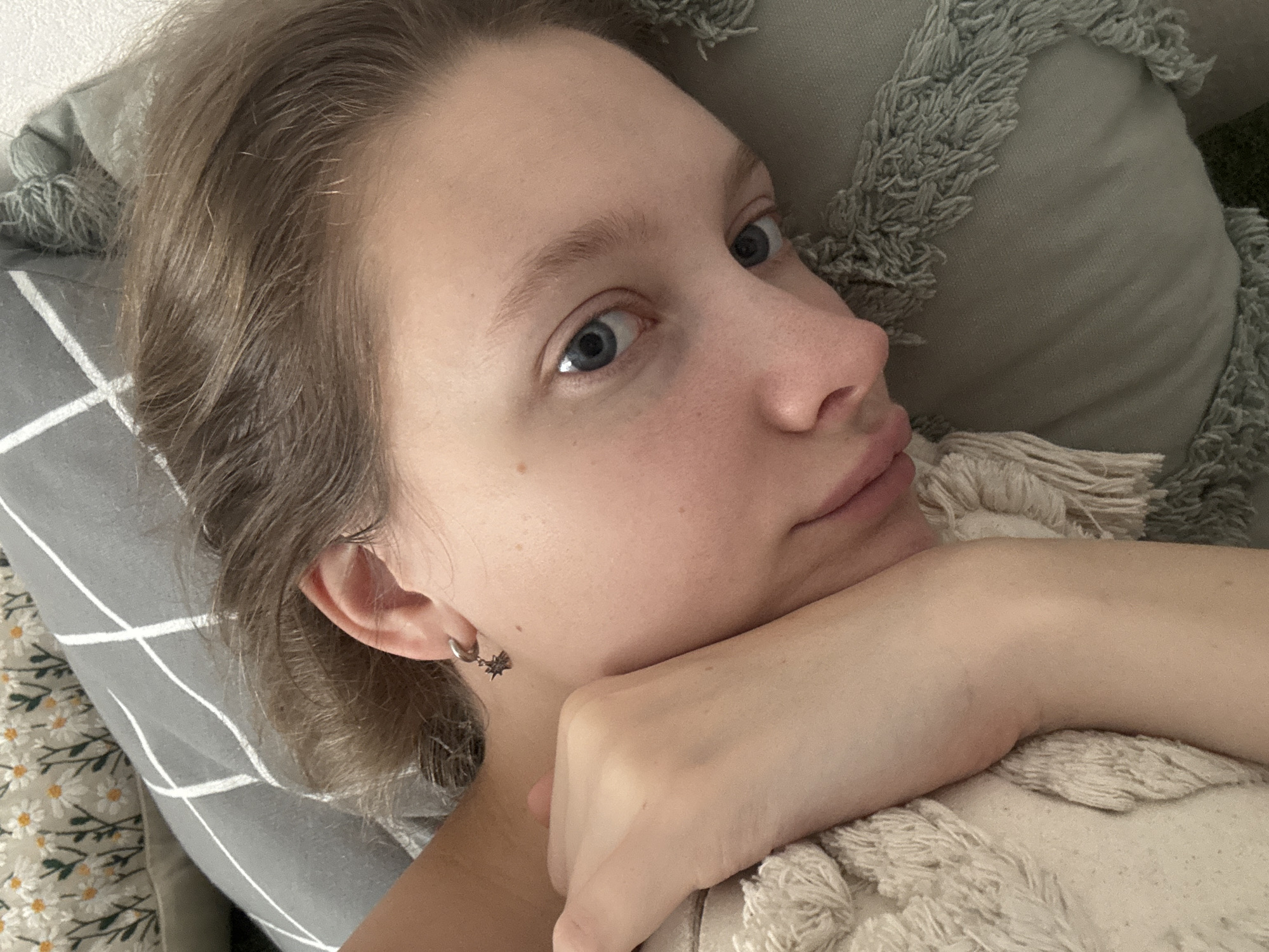The image is a close-up, low-angle selfie of a young person with fair skin and blue eyes, lying on a pillow. Their hair is light brown and appears to be slightly disheveled. The person is wearing a small, silver earring with a star-shaped charm. Their arm is resting on their cheek, and their eyes are looking upwards and to the right, giving a contemplative or perhaps surprised expression. The background consists of textured pillows. One pillow is grey with a white grid pattern, and another is a lighter, cream-colored pillow with a fringe. The lighting suggests it might be daytime, possibly indoors. The overall mood is relaxed and personal, typical of a casual selfie. There is no visible text in the image. The provided location context of Izhevsk, Russia, does not offer any direct visual cues within the image itself.
