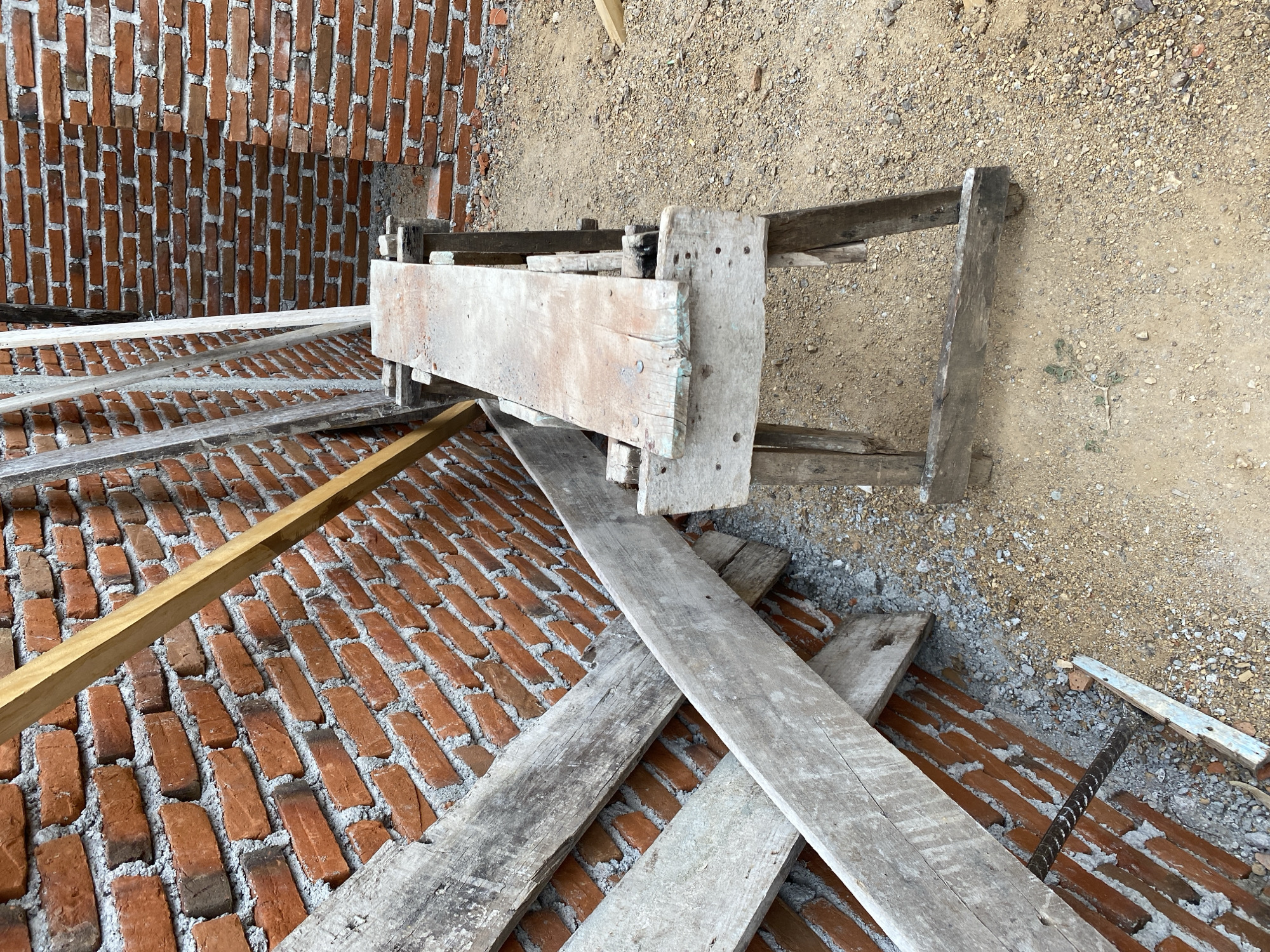 The image depicts an outdoor construction site in Banda Aceh, Indonesia. The foreground features a wooden sawhorse with a worn plank on top, resting on sandy ground. Behind the sawhorse, a partially constructed brick wall is visible, with several wooden planks leaning against it. Further in the background, more brick walls are apparent, suggesting the ongoing construction of a building. The scene is typical of a building project in progress, with raw materials and temporary structures in place.