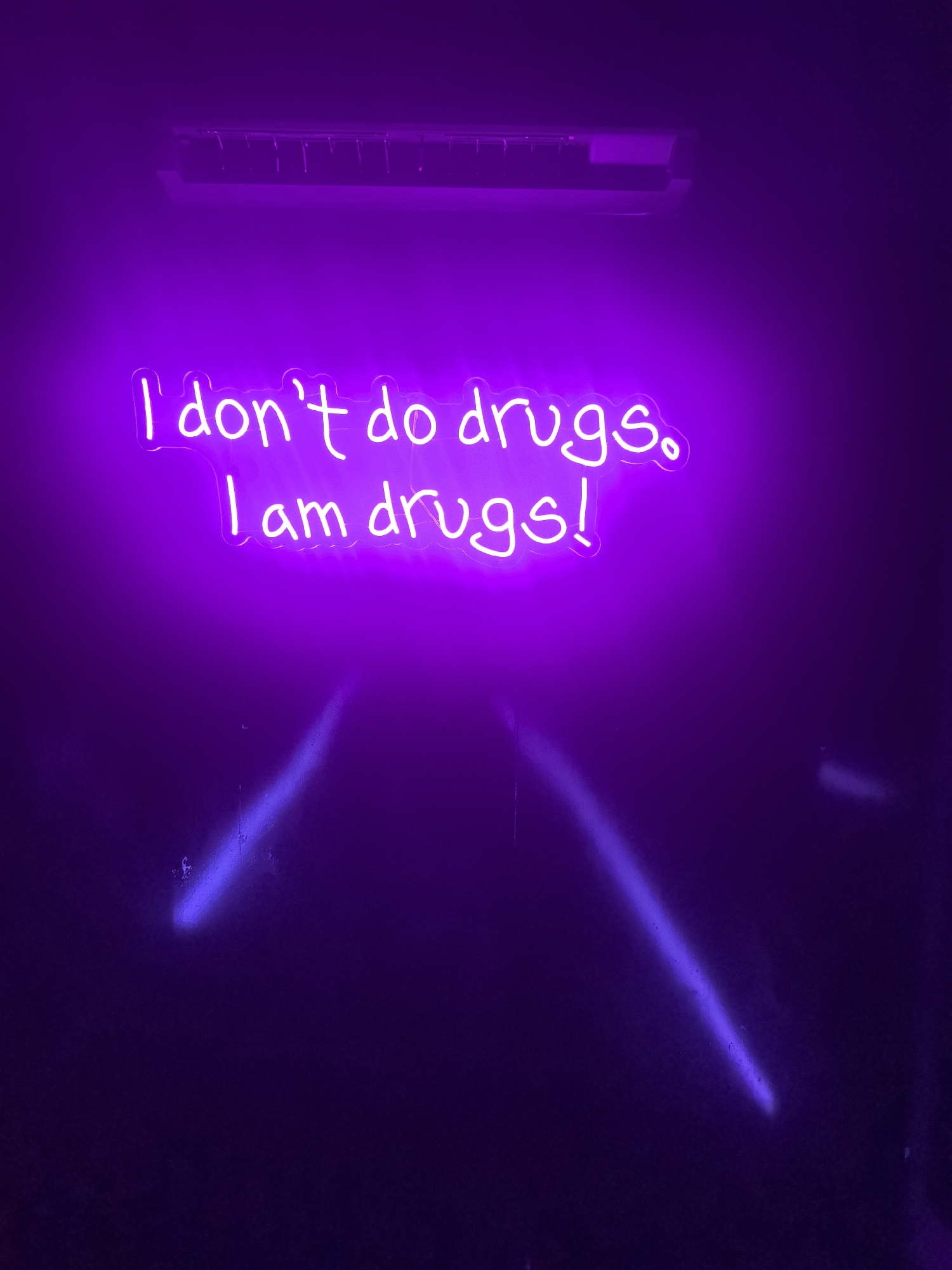 The image depicts a dark environment illuminated by a vibrant purple neon sign that reads "I don't do drugs, I am drugs!". The sign is positioned against a dark wall with some imperfections. Above the sign, there's a horizontal structure, possibly an air conditioning unit. Two streaks of purple light emanate from the bottom of the sign, adding to the overall dramatic effect. There's no information in the image to place it in Stara Zagora, Bulgaria or any other location.