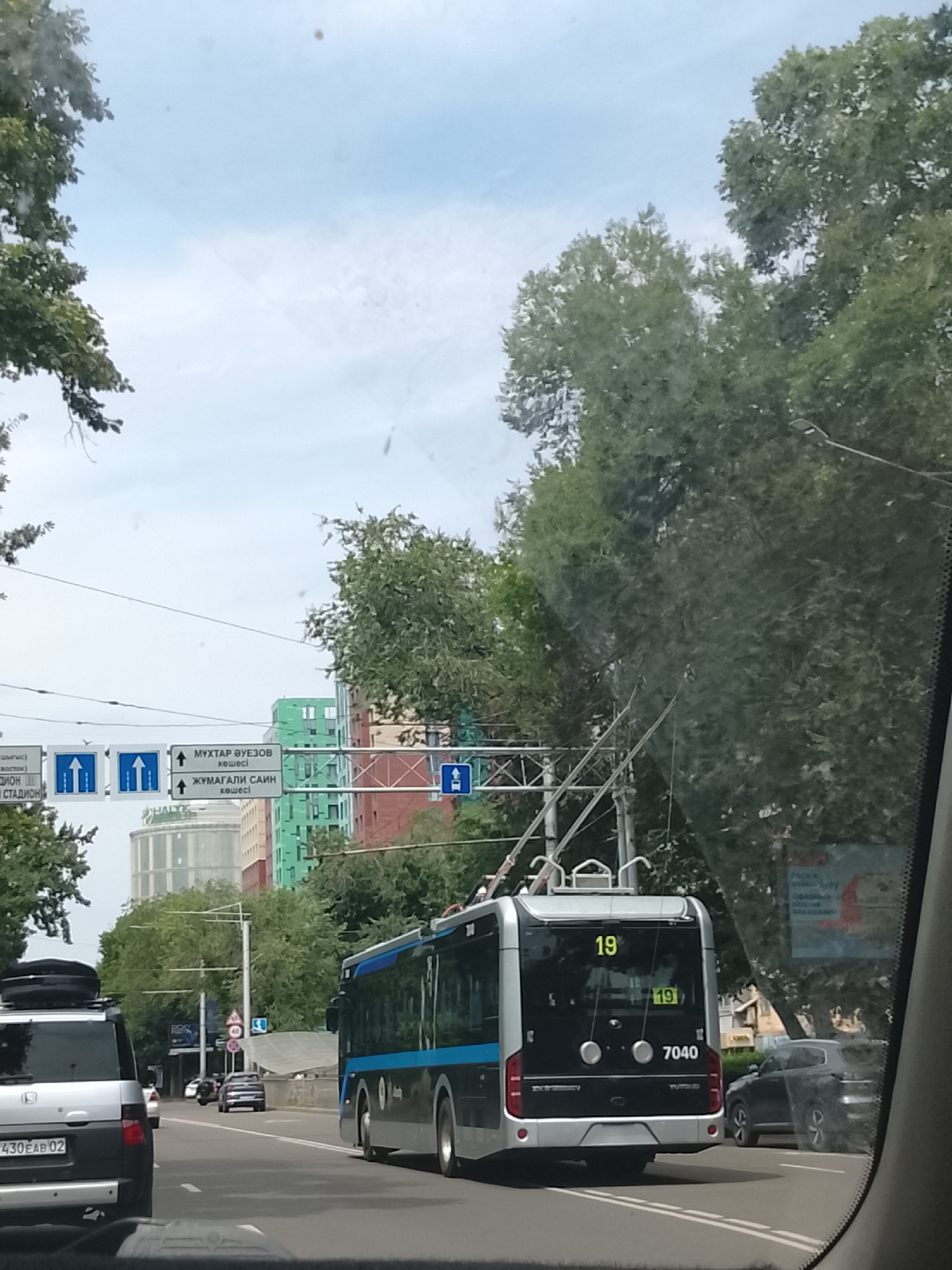 The image captures a street scene in Almaty, Kazakhstan, from the perspective of a vehicle's interior. A silver SUV with license plate 430EAB02 is visible in the foreground, its rear facing the camera. To its right, a black trolleybus with the number 19 displayed prominently on its rear and side travels down the road. The trolleybus has the number 7040 printed on its back. In the background, multiple modern buildings in various colors, including green and red, rise above a line of trees. Road signs are also visible, indicating directions to "МҰХТАР ӘУЕЗОВ" and "ЖҰМАҒАЛИ САИН." The sky is overcast.
