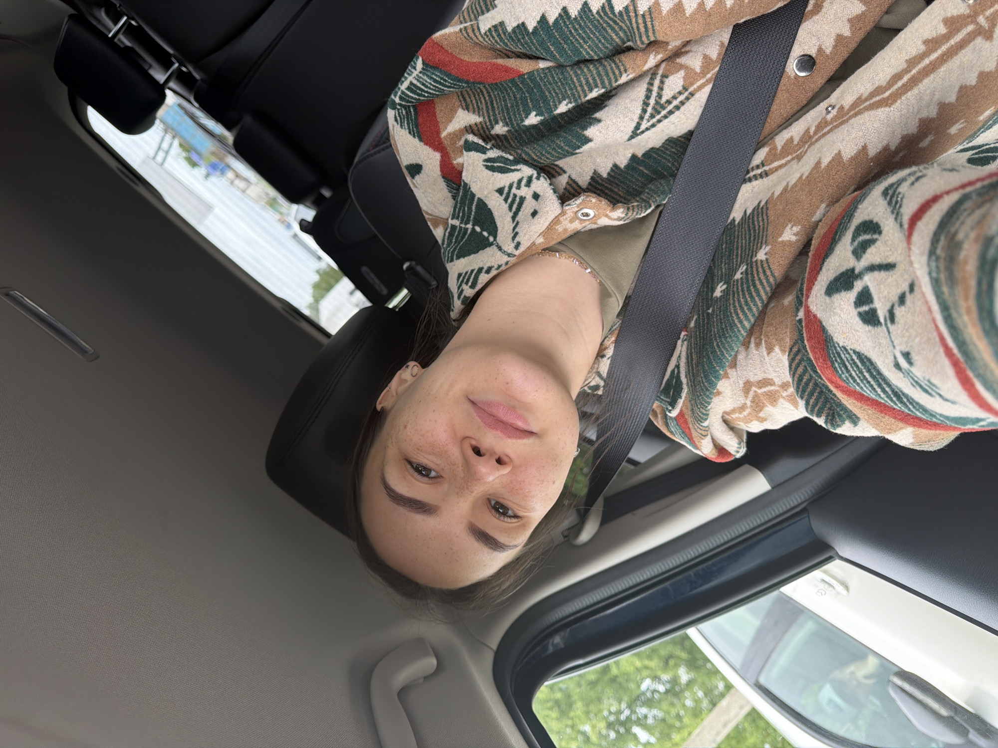 A woman with fair skin and freckles is sitting in the driver's seat of a car, wearing a patterned jacket and a seatbelt. The car's interior is visible, including the headrest of the seat behind her, the car door, and the roof. Outside the car window, there are trees and buildings, with a bright, overcast sky. The location appears to be in a residential or suburban area, possibly Chaikovsky, Russia.