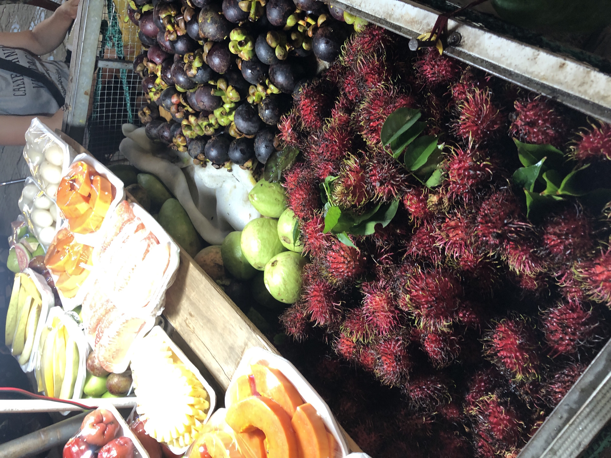 This media file depicts a vibrant fruit stall in Đà Nẵng, Vietnam. The scene is an overhead shot, showcasing an abundance of fresh fruits arranged for sale. In the foreground, several clear plastic containers hold sliced fruits, including bright orange papaya and a white fruit, possibly pineapple, sliced into florets. Other containers feature whole fruits like starfruit and what appear to be rose apples.
Dominating the right side of the frame are large piles of rambutan, recognizable by their bright red, spiky exterior. To the left of the rambutan, a substantial mound of mangosteens, with their deep purple rind and green calyx, are visible. In the background, a person's arm and shoulder are visible, wearing a grey t-shirt with faded text. The setting appears to be an outdoor market or a fruit stand, given the rustic wooden crates and metal shelving. The lighting suggests it could be daytime, possibly late morning or afternoon, with natural light illuminating the colorful produce. The overall impression is one of a bustling market scene, rich with tropical fruits. There is no visible text in the image, apart from what might be on the person's t-shirt, which is too blurry to read.