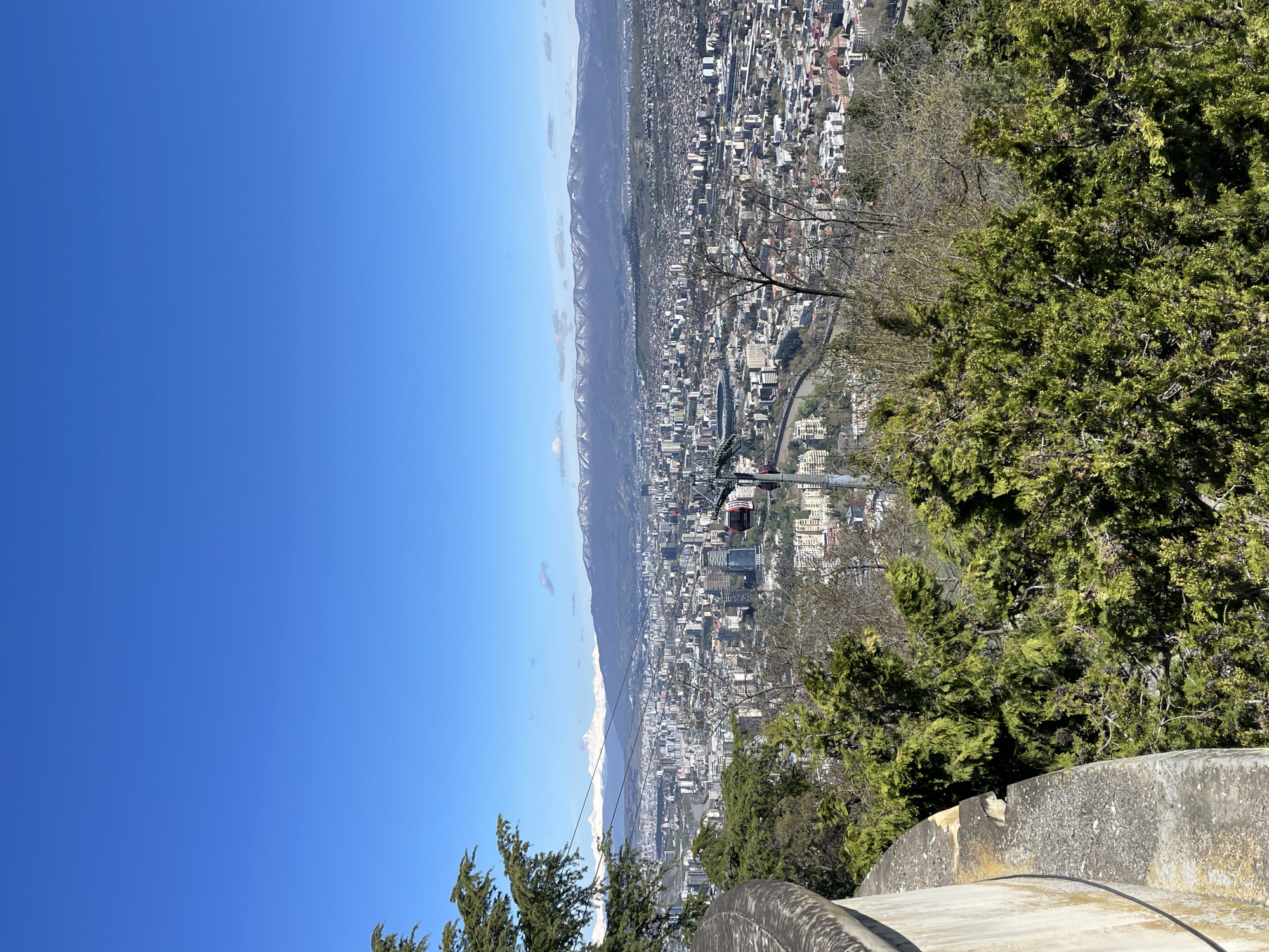 The image shows an elevated view of the city of Tbilisi, Georgia, on a clear, sunny day. In the foreground, there are evergreen trees and a portion of a concrete or stone structure, possibly a viewing platform or building. A cable car with a red cabin is visible, suspended over the city. The city itself is densely built, with a mix of high-rise and low-rise buildings spreading across the landscape. In the background, there are mountains. The sky is a clear, bright blue.