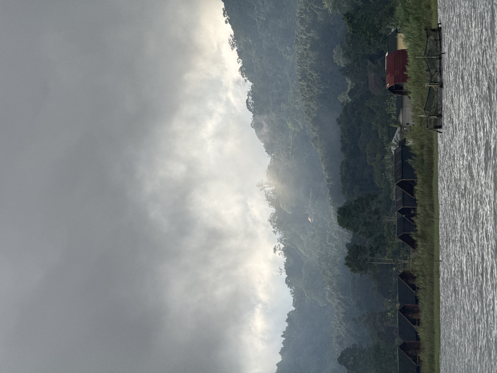 The image shows a lakeside view in Baturiti, Indonesia. Mountains are on one side, partially obscured by fog and clouds. A row of small, dark-colored buildings is situated along the grassy shoreline of the lake, with additional structures further along the water. The lake itself has a rippled surface. The scene is overcast with muted lighting.