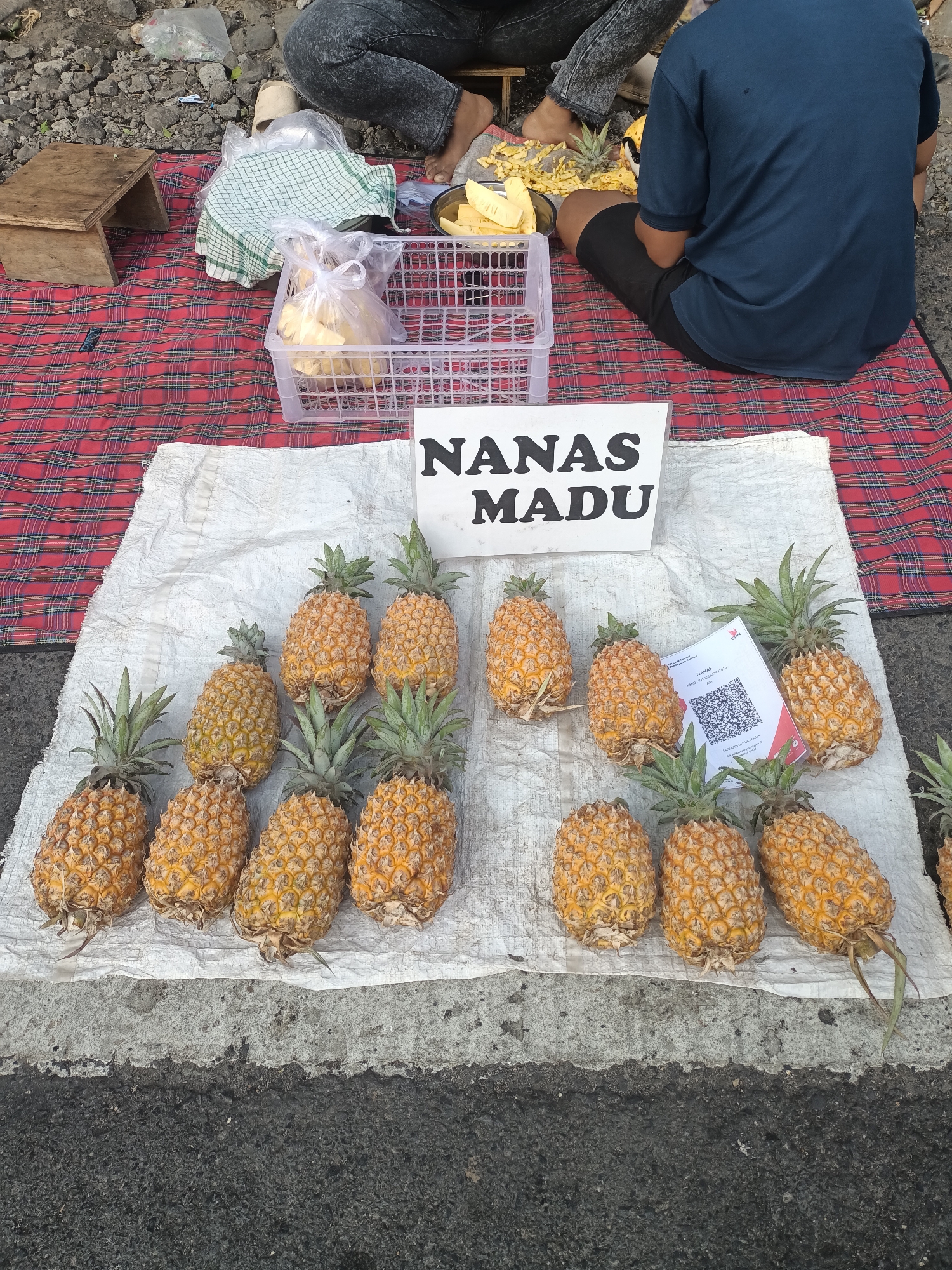 A market stall in Pare, Indonesia, displays a selection of ripe pineapples for sale. The pineapples are arranged on a white cloth spread over a grey concrete surface, with a red and black checkered blanket visible in the background. Several people are seated behind the stall. One person, wearing a blue t-shirt and black shorts, is partially visible from the back, while another person, wearing dark jeans, is seated on the blanket. A small wooden stool and a plastic basket filled with pineapples are also present. A sign with "NANAS MADU" written on it, indicating "Honey Pineapple," is placed prominently in front of the pineapples. A QR code is also visible on a small white card near some of the pineapples. The scene suggests a typical roadside market offering fresh produce.