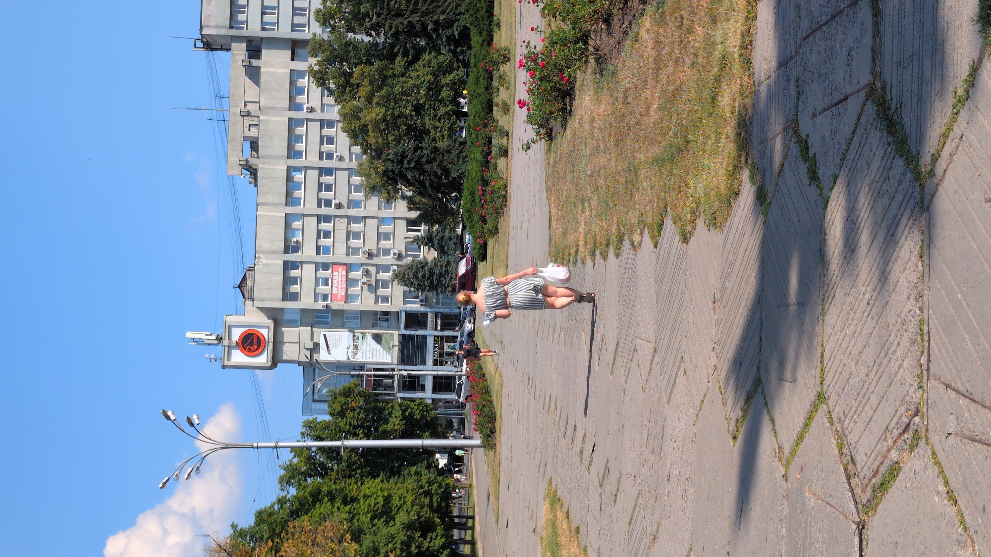 In Poltava, Ukraine, a woman in a striped dress walks away from the camera down a paved pathway in a park. She carries a white plastic bag in her right hand. The pathway is bordered by a dry, grassy area and a flower bed with red roses. In the background, a multi-story building with many windows is visible, along with trees and a street with cars. The sky is clear and blue.