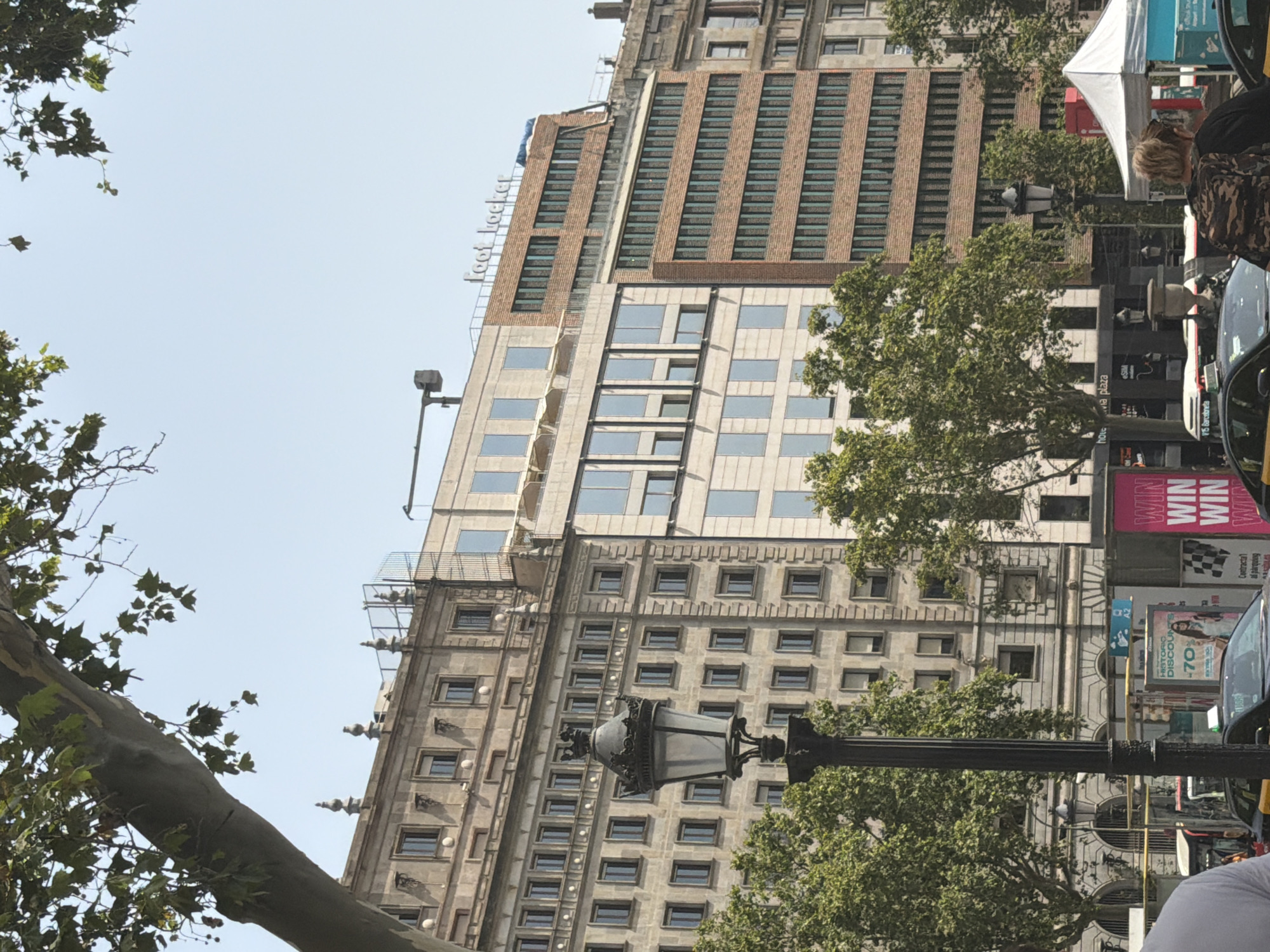 The image shows a street scene in Barcelona, Spain, on a sunny day with a clear sky. In the foreground, a black lamppost with a decorative crown-like top is prominent. Behind it, several buildings of varying architectural styles are visible. The leftmost building is a light-colored, multi-story structure with numerous windows and ornate detailing. To its right, a more modern, light-colored building with a facade of rectangular windows is present, topped by a "Foot Locker" sign in white lettering. Adjacent to that is a brick building with a pattern of dark, recessed windows. Further to the right, another older building with stone detailing can be seen. Green trees are scattered throughout the scene, some obscuring parts of the buildings. In the lower portion of the image, yellow taxis, part of a white bus, and various signs and advertisements are visible, suggesting a busy urban environment. People are present, including a person wearing a camouflage backpack in the mid-ground and a person's shoulder in the left foreground. A white tent structure is also visible on the right side.